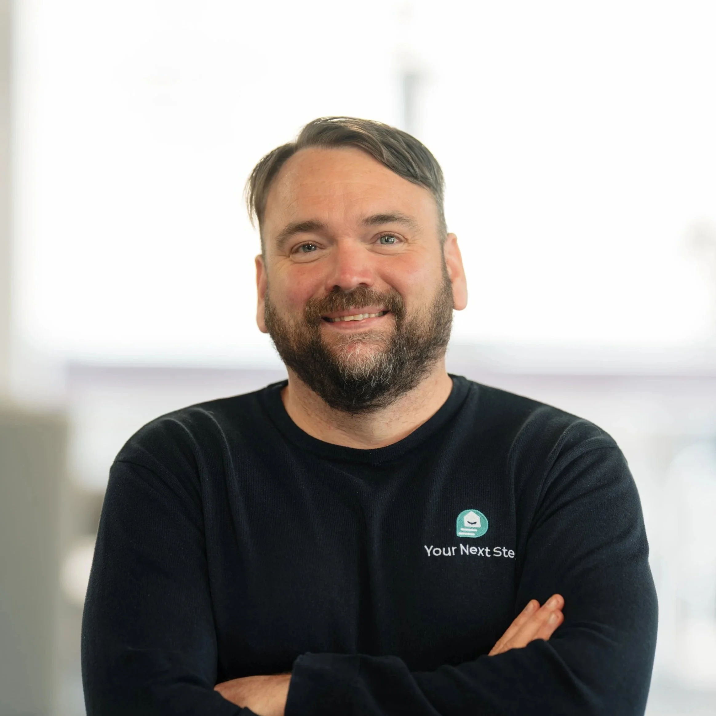 A man with short hair and a beard, smiling, wearing a navy blue polo shirt with a logo and the text 'Your Next Step' on it, sitting against a light mint green background.