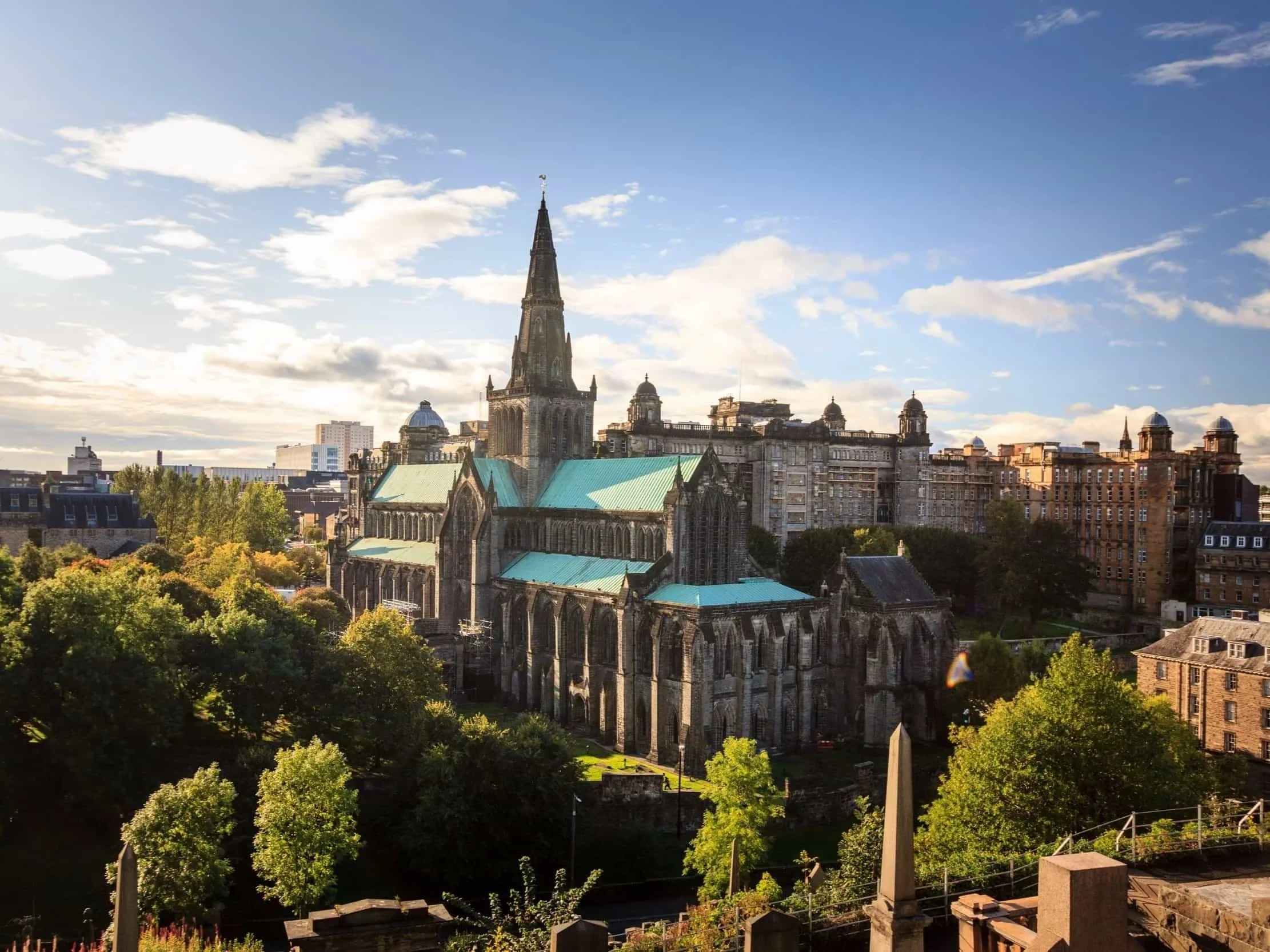 A cityscape with a prominent historic church featuring a tall steeple, surrounded by various buildings and green trees, under a partly cloudy sky