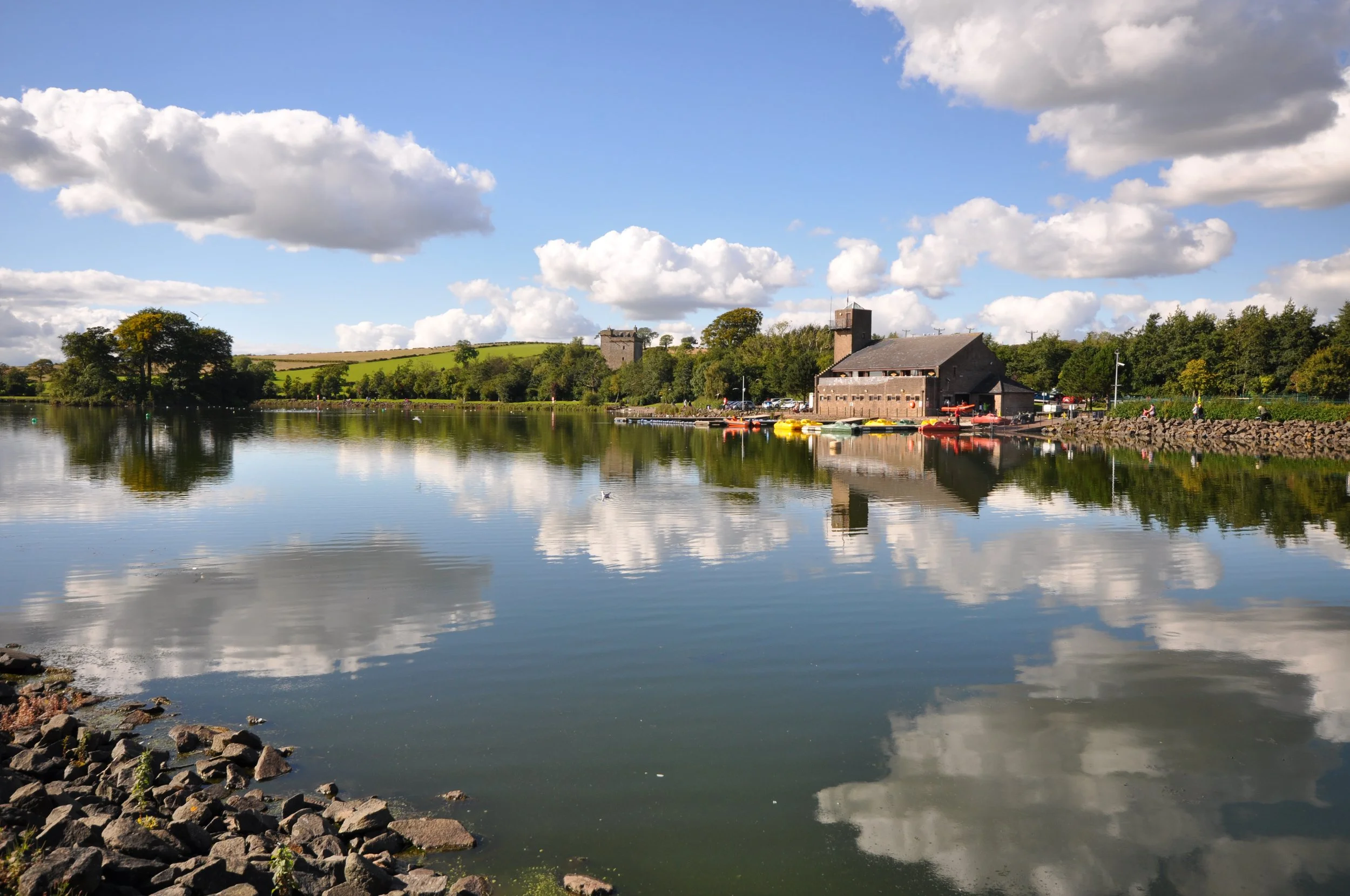 A peaceful lakeside scene with boats docked along the pier, a stone building near the shore, lush green trees, a grassy hill, and a blue sky with scattered clouds reflected in the calm water.