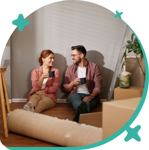 A man and woman sitting on the floor inside, holding mugs and smiling at each other, next to a large rolled-up rug, with a plant and a staircase in the background.