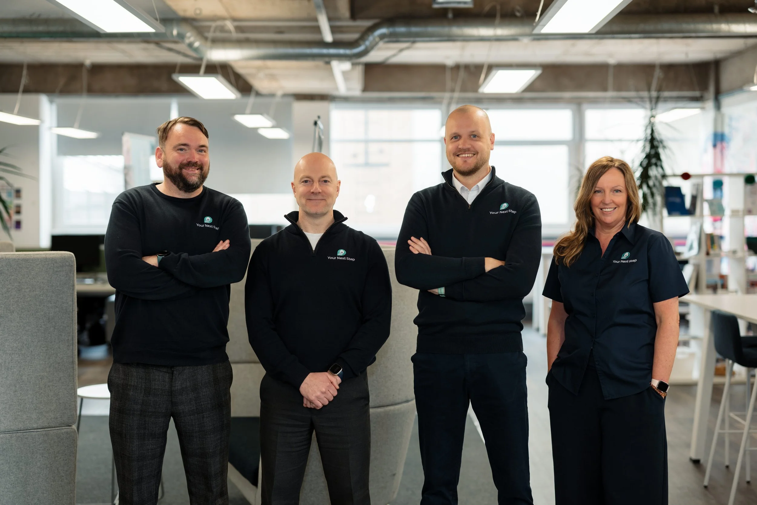 Four team members standing in an office, smiling at the camera, wearing matching black shirts with a logo and slogan, with an open workspace background.
