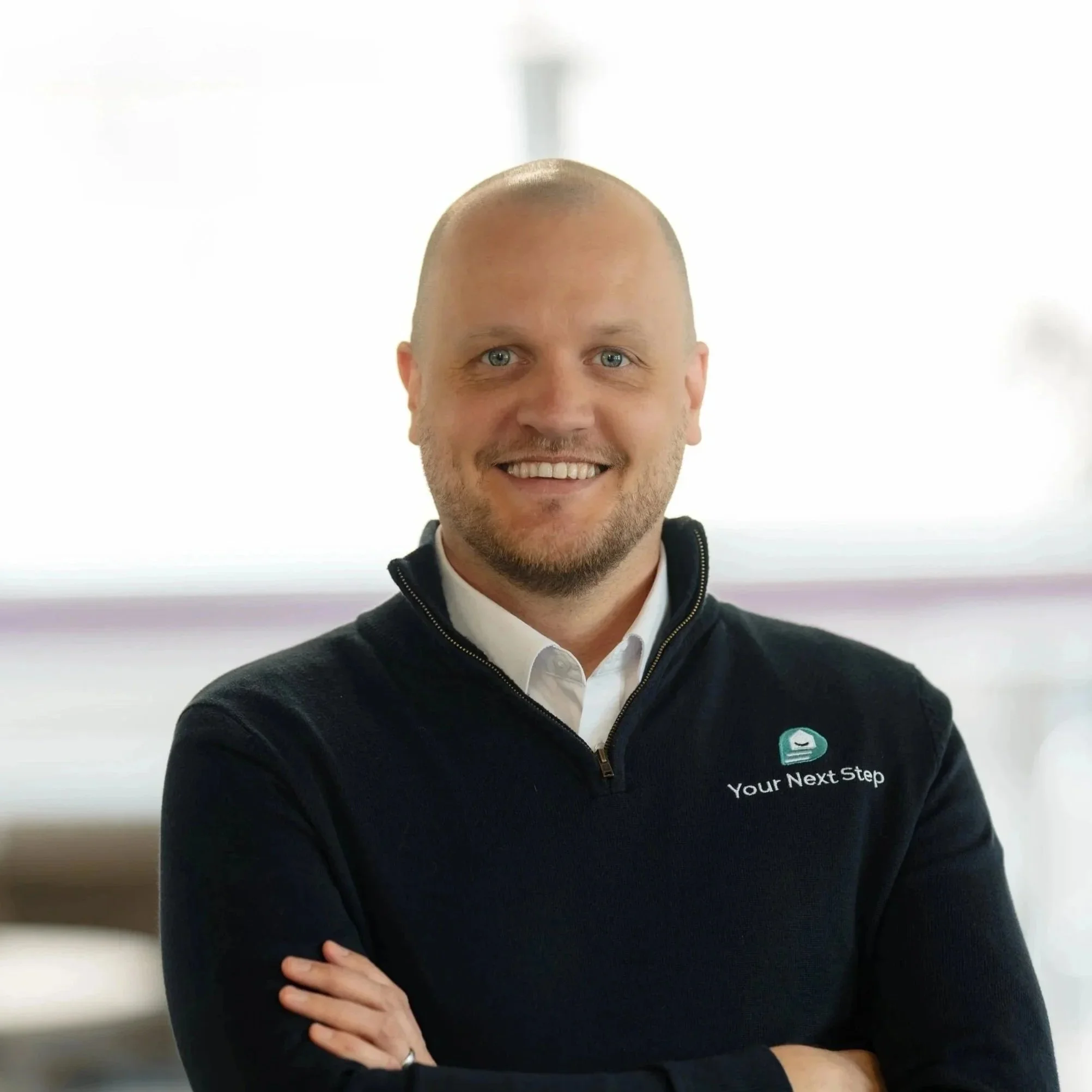 A professional man with a beard and short hair, wearing a dark blazer and white shirt, smiling with arms crossed, posed against a light teal background.