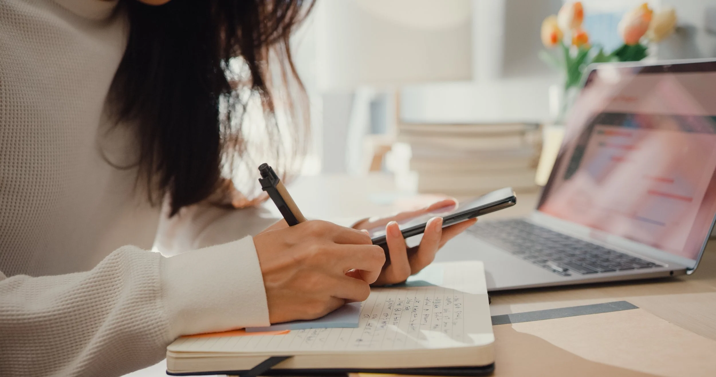 A person writes in a notebook with a pen while working on a laptop, with a tablet in hand nearby, in a bright, organized workspace with a vase of flowers and stacked papers.