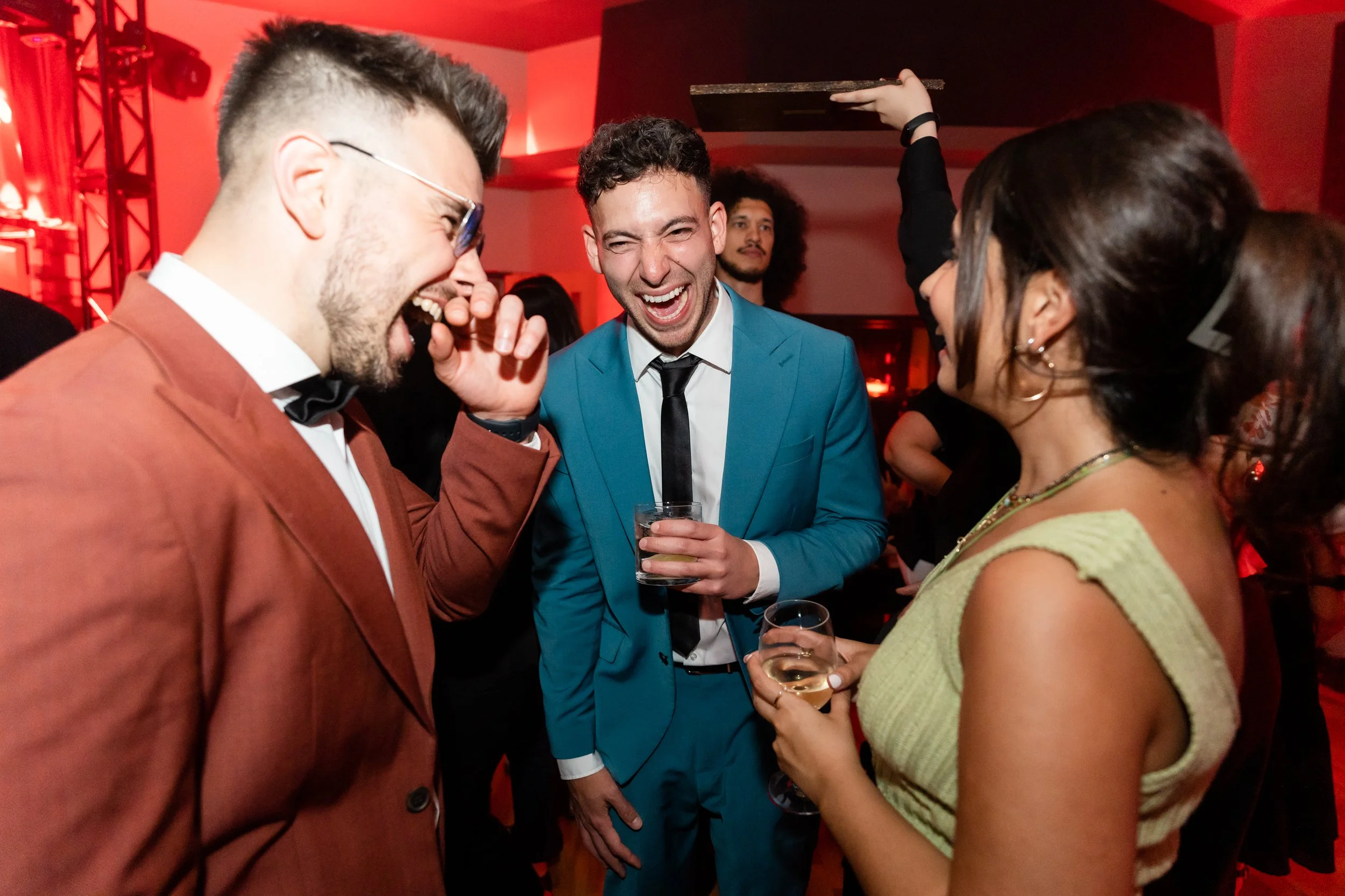 People laughing and talking at a social event, dressed in formal attire, with drinks in their hands.