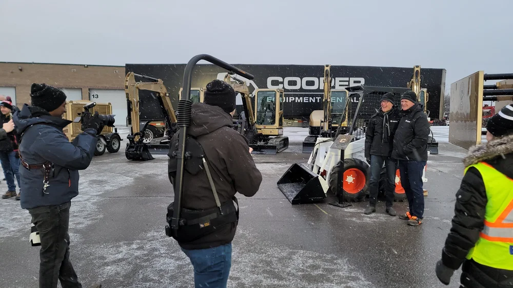 People and equipment at a construction rental site with photographers taking pictures in a snowy setting.
