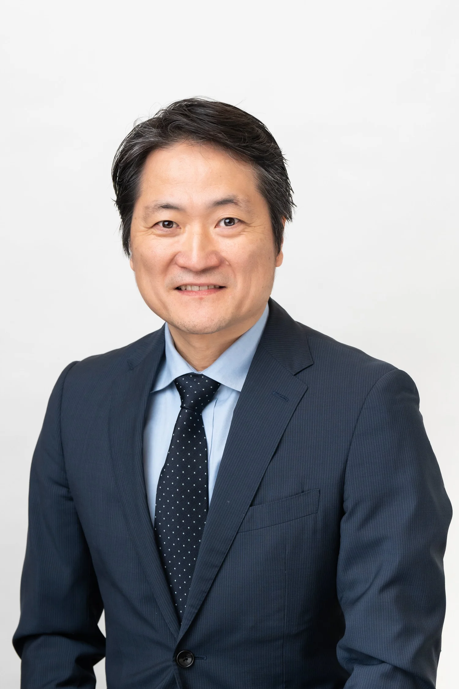 A man in a dark suit and a light blue shirt, wearing a polka dot tie, smiling against a plain white background.