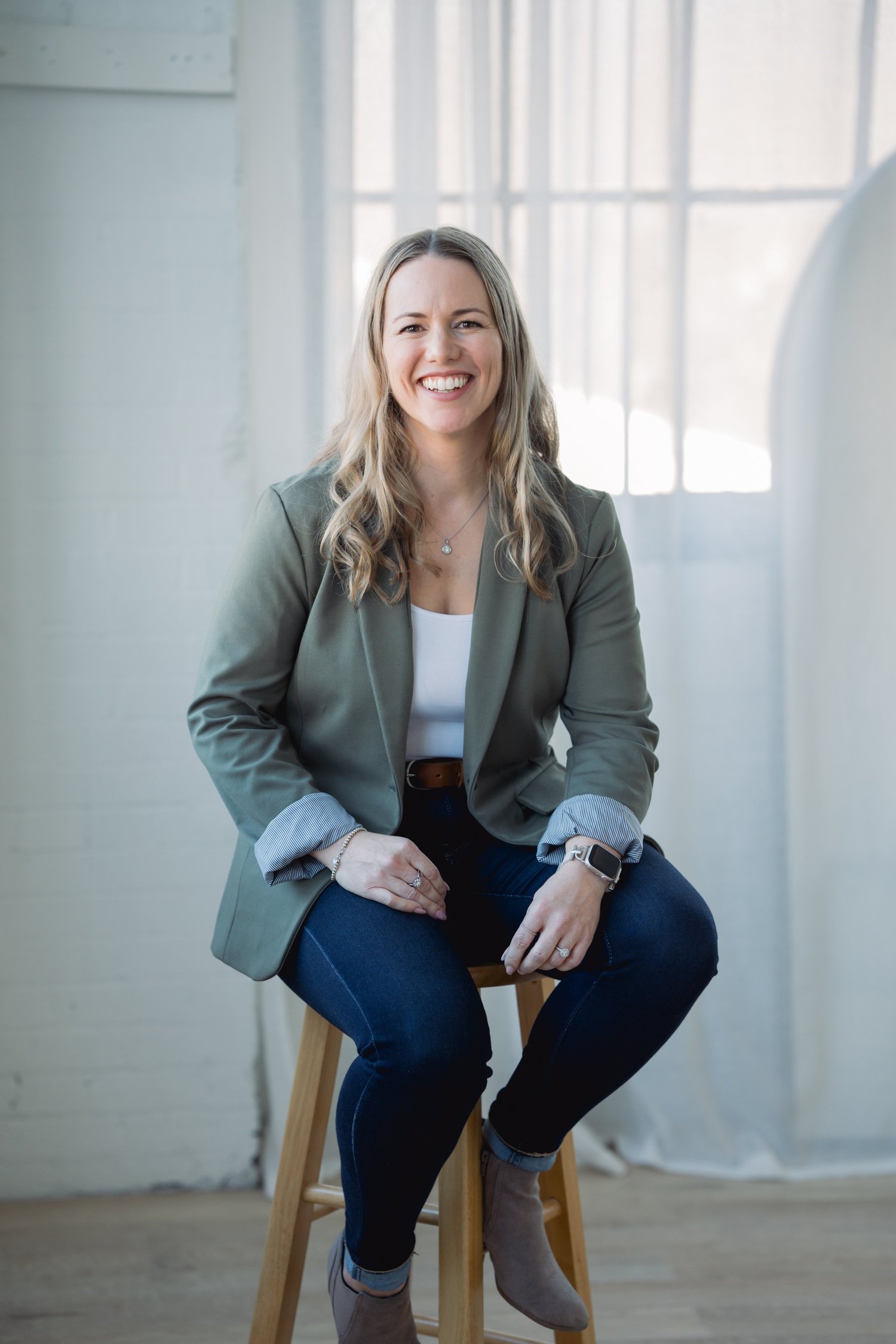 Smiling woman sitting on a wooden stool, wearing a green blazer, white top, and jeans in a bright indoor setting.