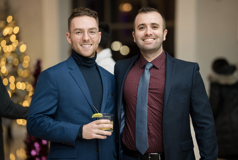 Two men in formal attire standing together, one holding a drink with a lime, in a festive setting with a blurred background of lights and a decorated tree.