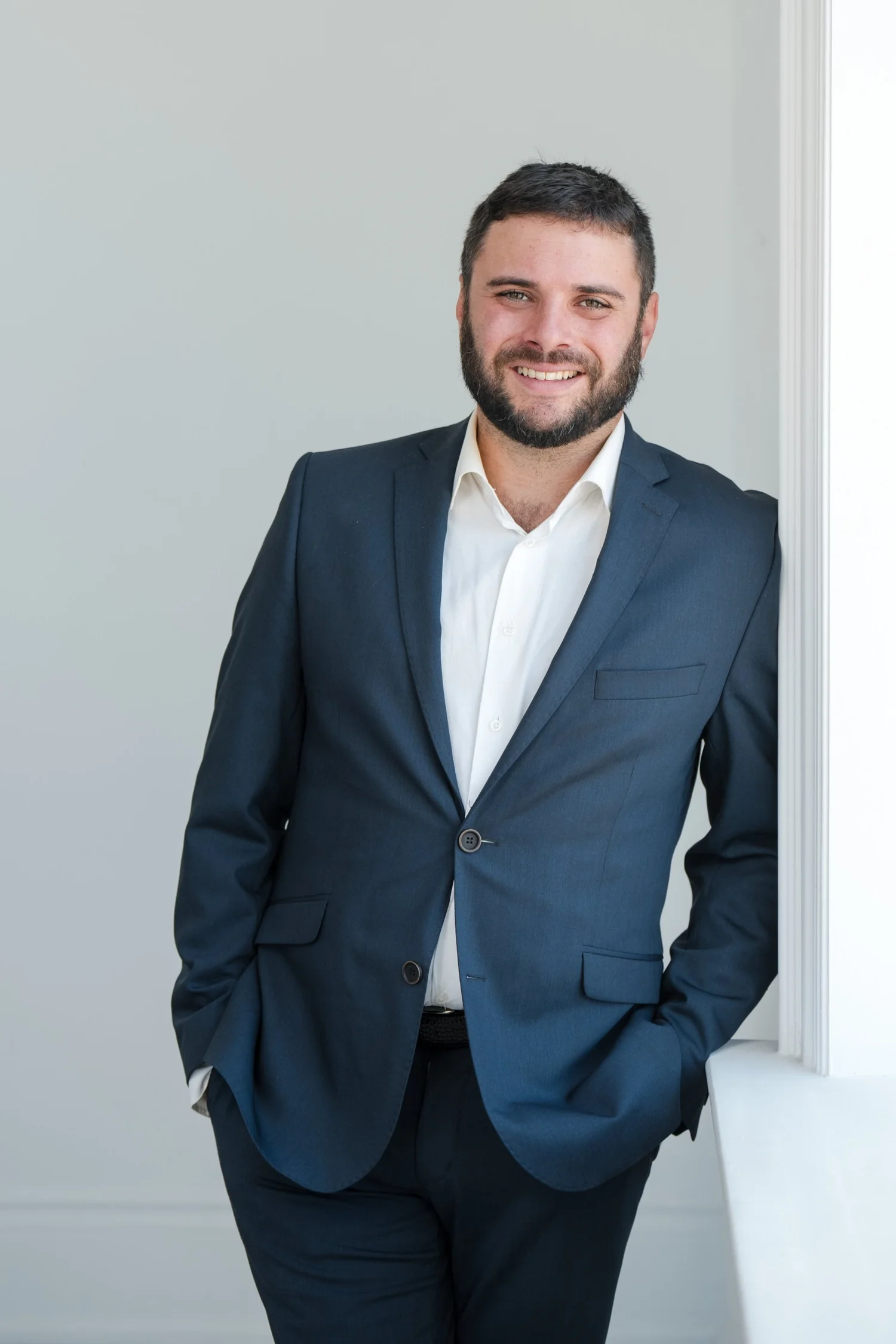 Man in a dark suit with a white shirt leaning against a white wall, smiling.