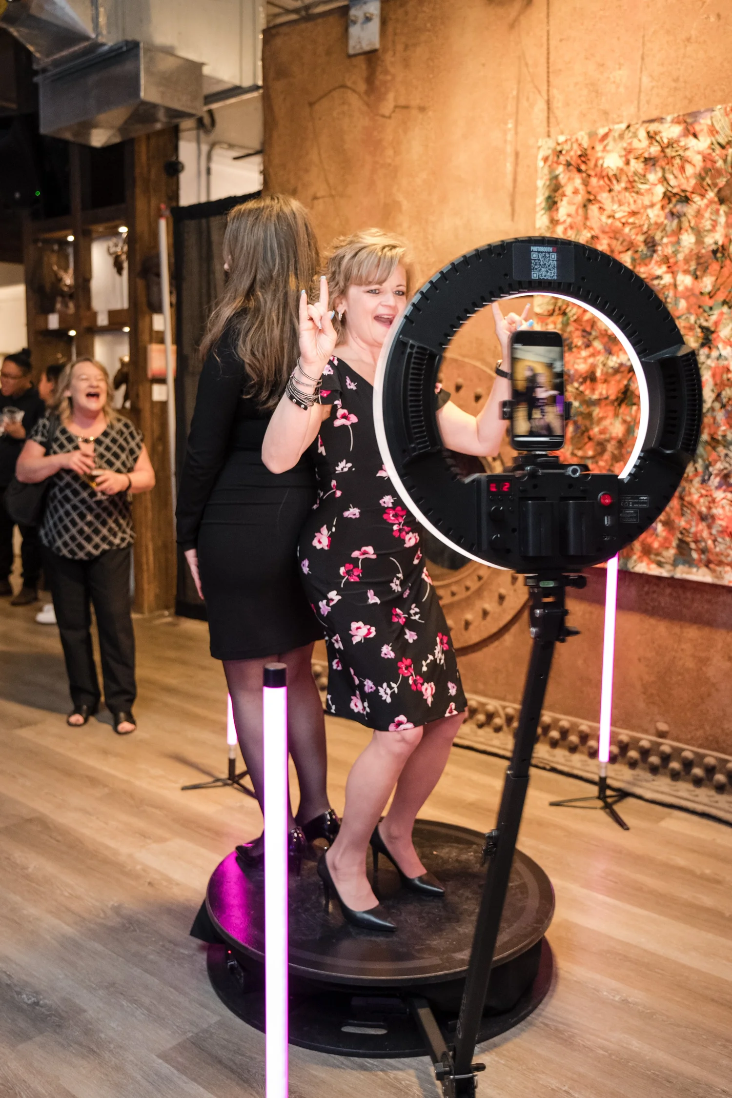 Two women standing on a rotating platform surrounded by LED lights, taking a selfie with a smartphone mounted on a ring light tripod. They are dressed in formal attire, and people in the background are watching and laughing.
