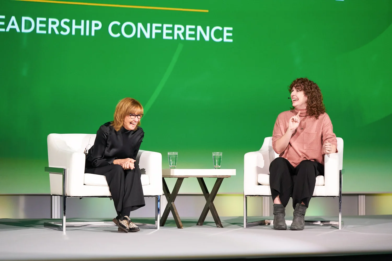 Two women seated on stage at a leadership conference, engaging in conversation with microphones, set against a green backdrop.