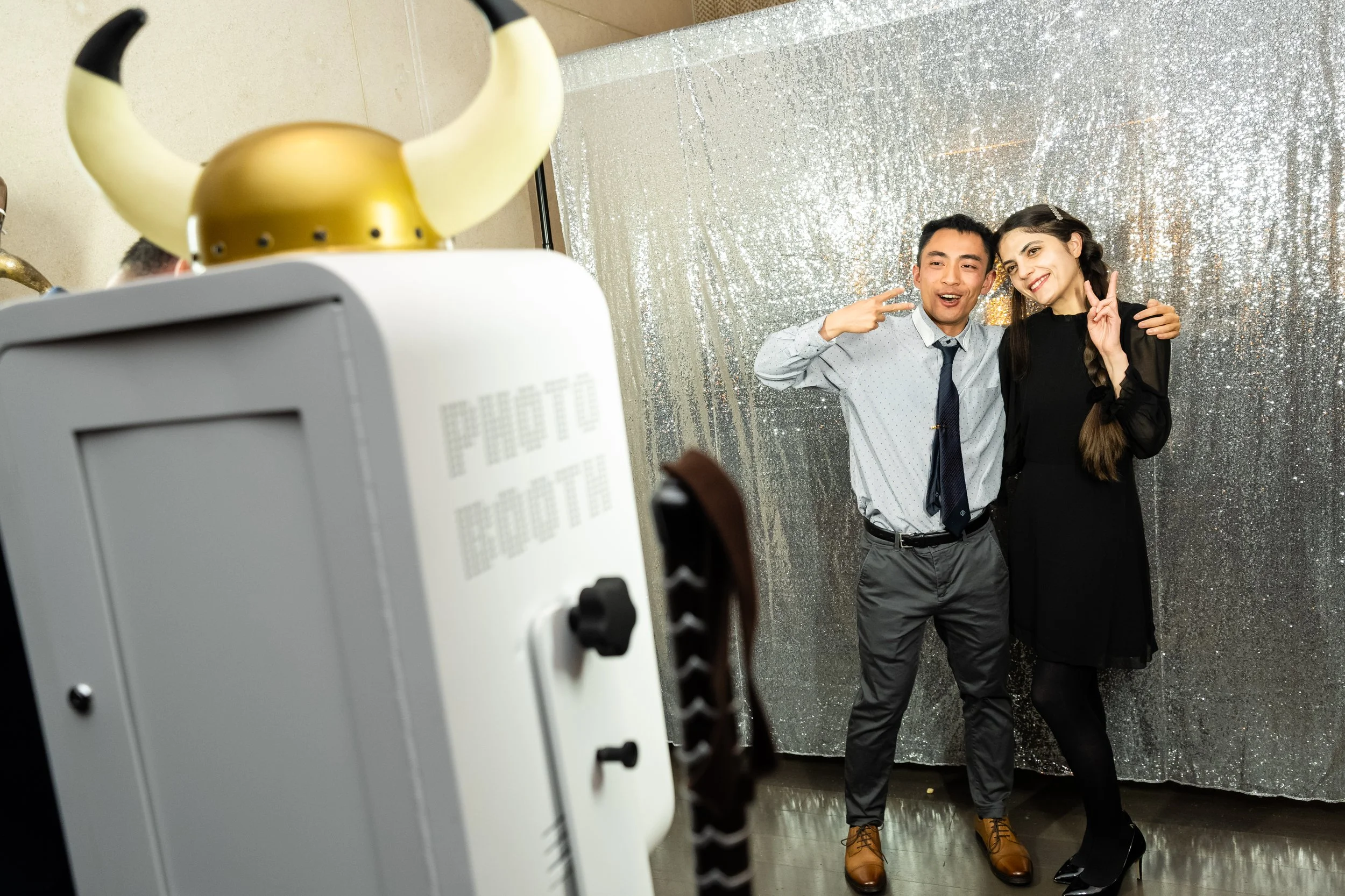 Two people posing for a photo booth against a shiny silver backdrop, with one making a peace sign and the other smiling. The photo booth features a horned helmet on top.