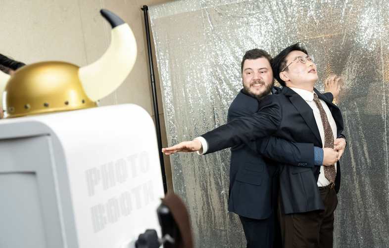 Two men in suits posing playfully in a photo booth with a Viking helmet prop in the foreground.