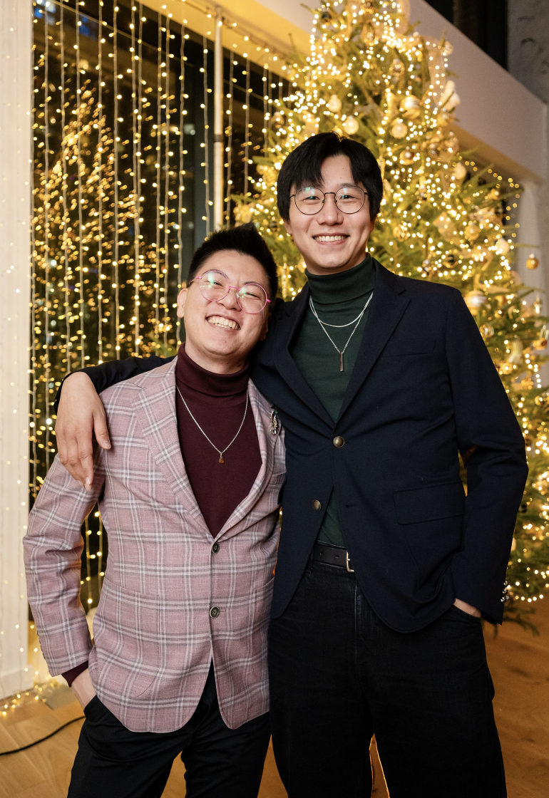 Two people standing in front of a decorated Christmas tree with lights. They are both wearing blazers and smiling at the camera, with one person having their arm around the other.