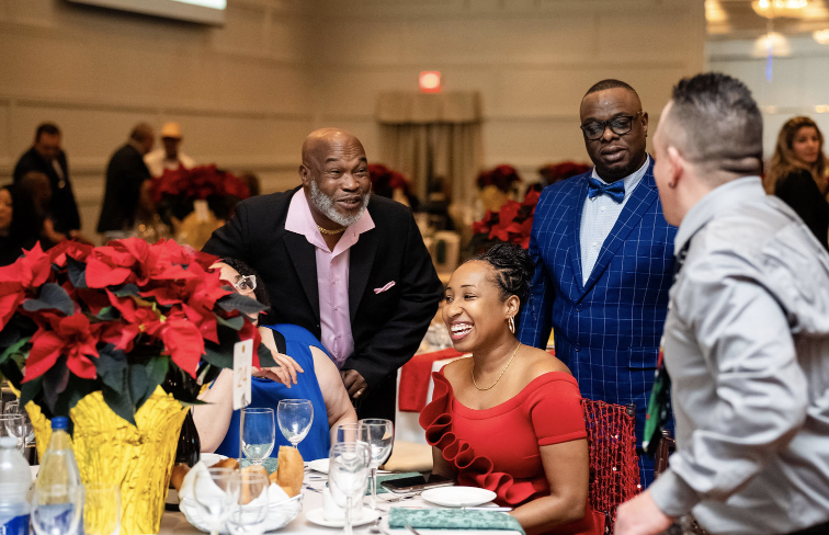 A group of people socializing at a banquet table during a formal event. The table is decorated with poinsettias and set with glassware and plates. The attendees are dressed in formal attire, smiling and conversing.