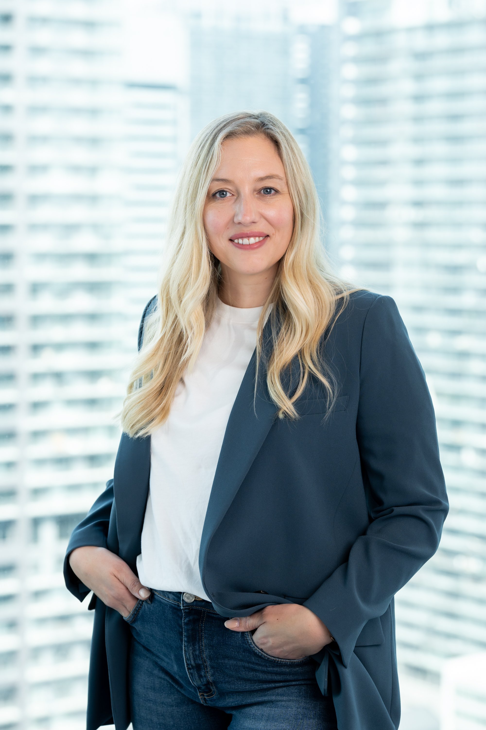 Woman with long blonde hair wearing a blazer and jeans, standing in front of a cityscape background.
