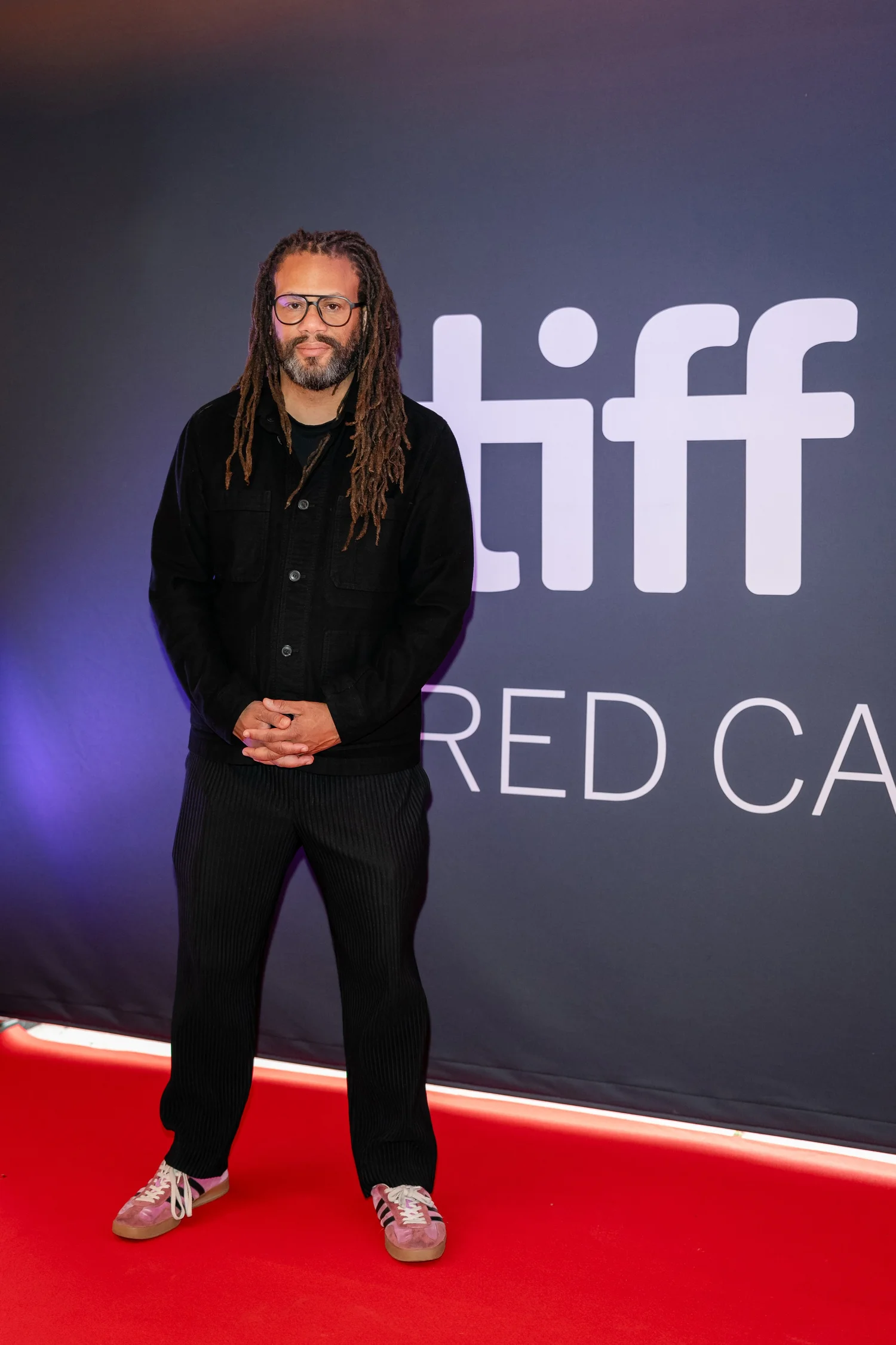 A person with long hair and glasses stands on a red carpet in front of a backdrop with 'tiff' and 'RED CAR' visible. They are wearing a black outfit and sneakers.