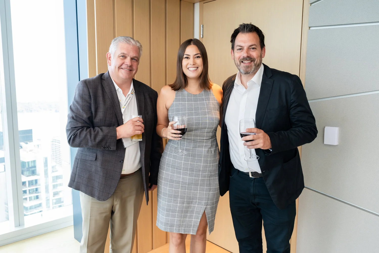 Three people smiling, standing together indoors, holding drinks, with a city view in the background.