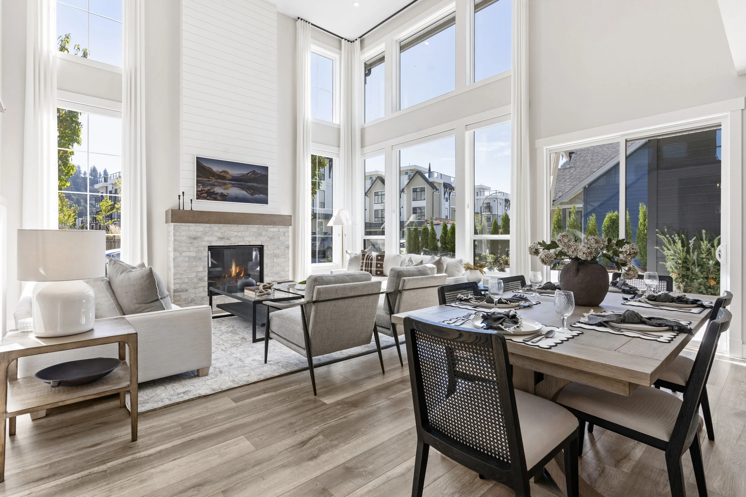 Show home featuring an open-concept living room, dining area, and kitchen with large windows, white walls, modern furnishings, and lots of natural light, including a sofa, armchairs, a fireplace, and a beautifully styled dining table.