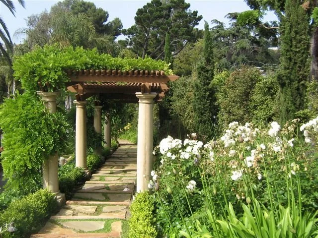 A garden pathway with stone steps leading to a wooden pergola supported by white columns, surrounded by lush green plants and white flowers.