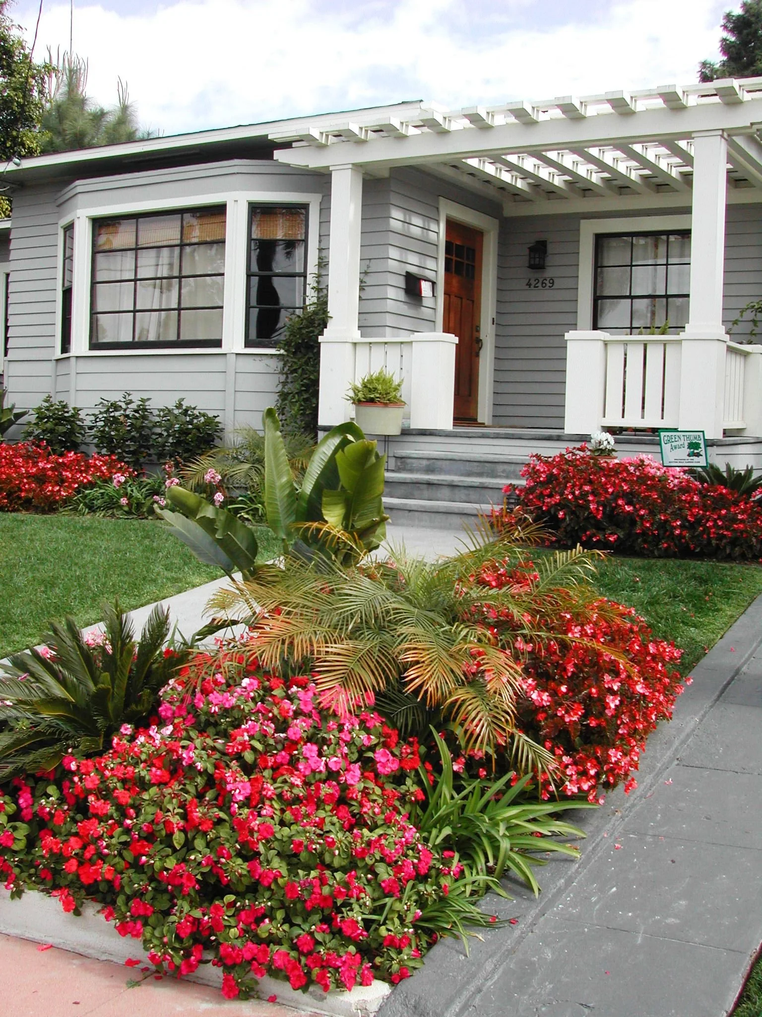 A house with gray siding and white trim, featuring a front porch with steps, a wooden front door, and large windows. The front yard is landscaped with colorful flowers, including red and pink blooms, and various green plants. There's a small sign on the sidewalk indicating a "Green Thumb Award."