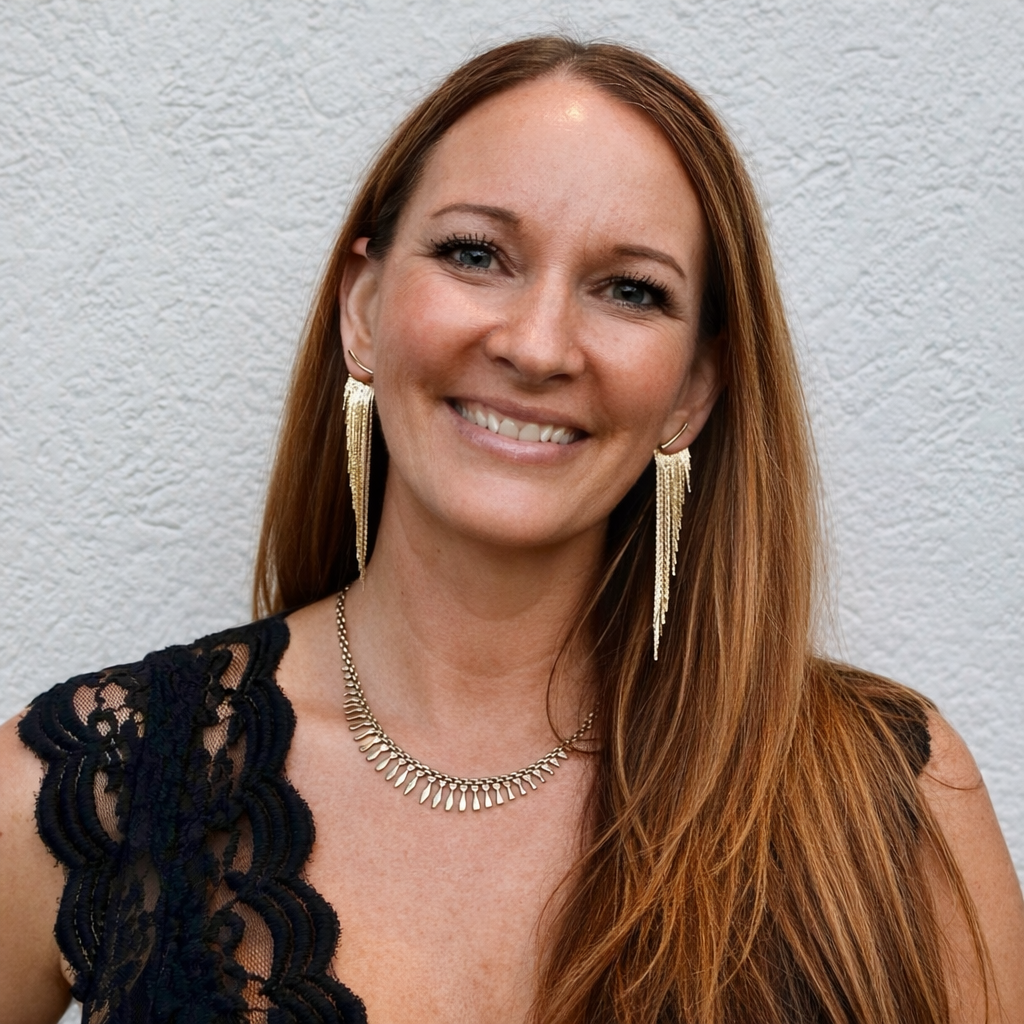 Close-up of a woman with long red hair, wearing a black lace top, gold dangling earrings, a necklace, and smiling against a textured white background.