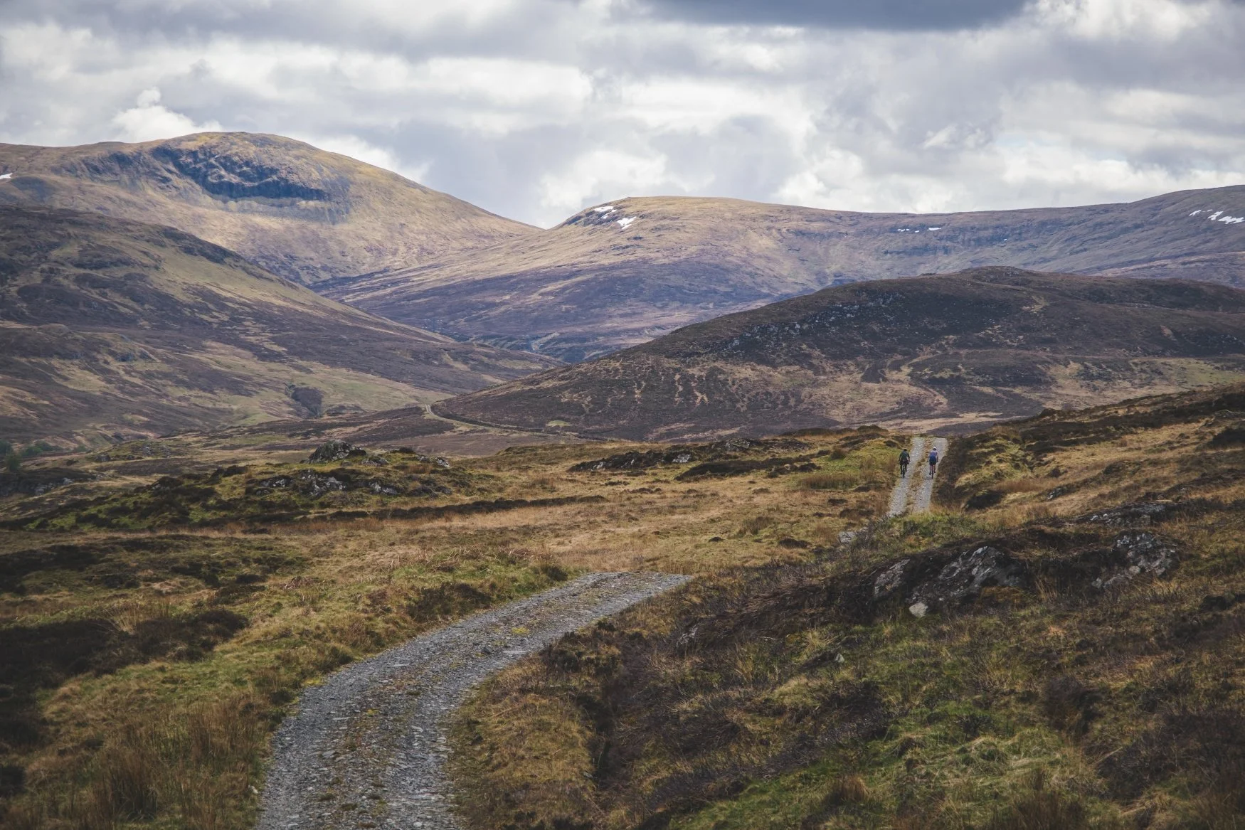 Two hikers walking on a narrow trail through grassy and rocky terrain in a mountainous landscape under a cloudy sky.