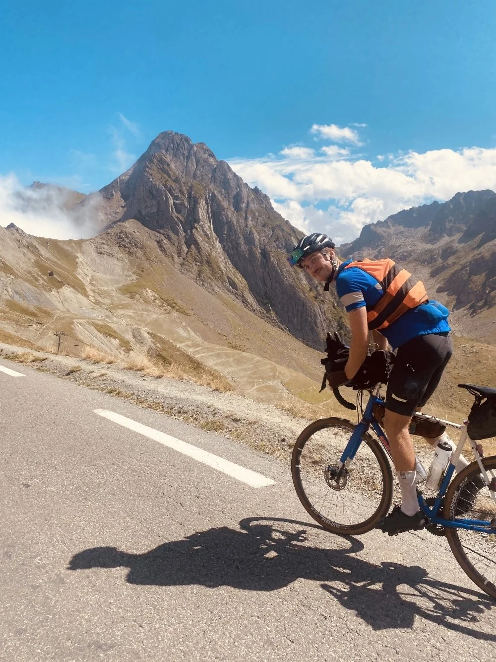 A male cyclist in a blue shirt, black shorts, and a helmet riding a mountain bike on a mountain road with rocky peaks and a partly cloudy sky in the background.