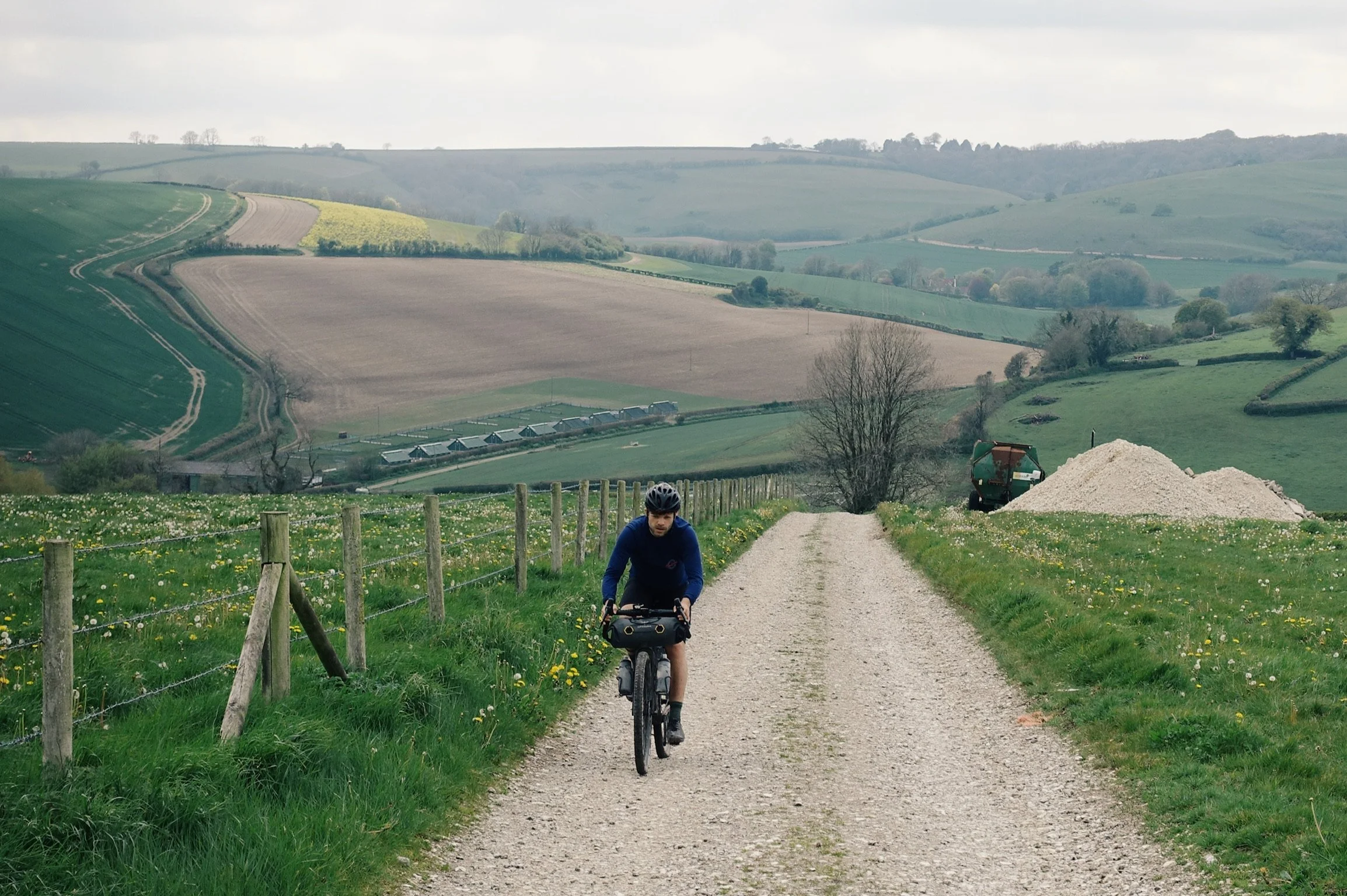 A person cycling on a gravel country road with green fields, wooden fence, and rolling hills in the background.