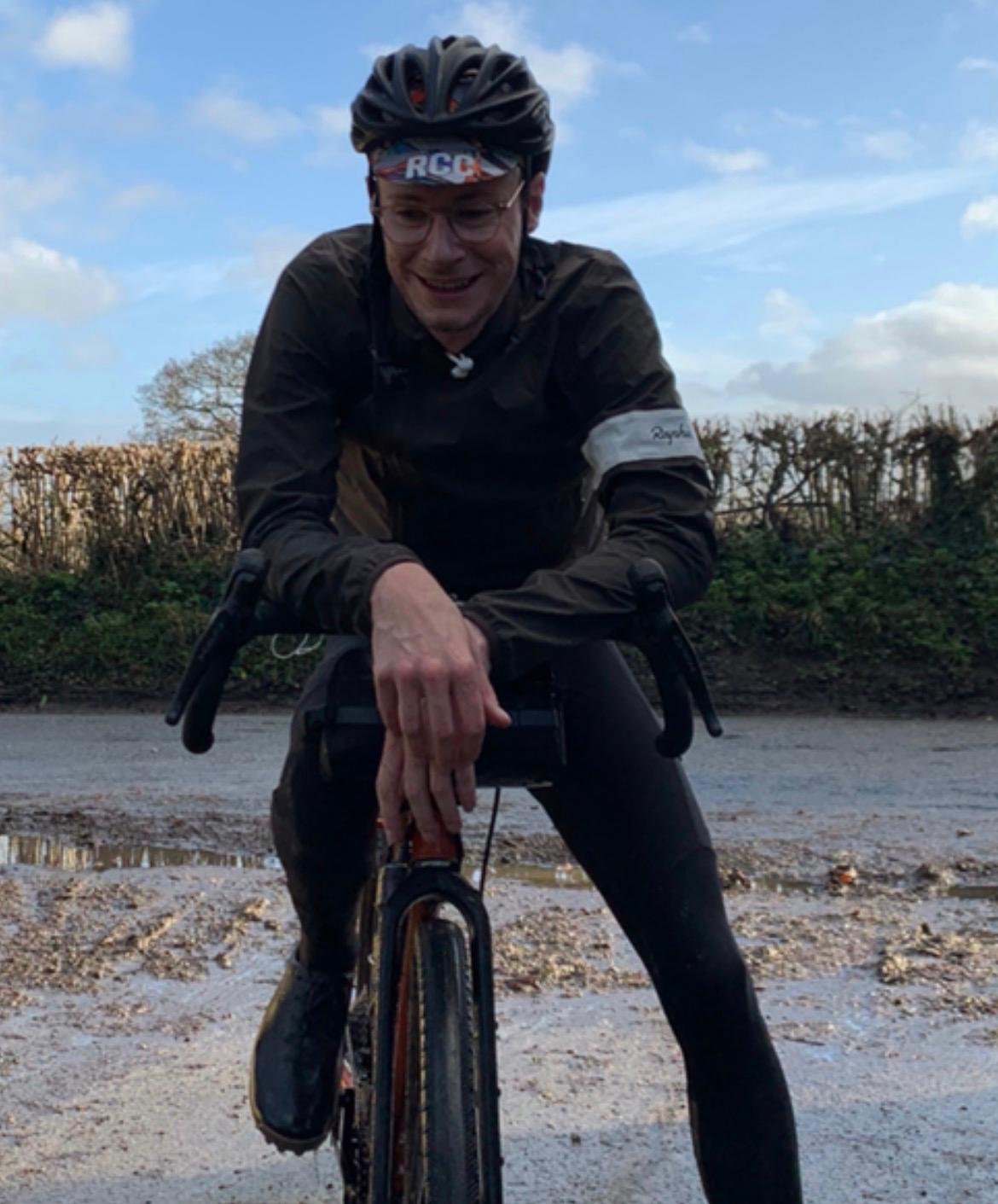 A man wearing a bicycle helmet, glasses, and athletic clothing, smiling while riding a bicycle outdoors, with trees and a fence in the background.