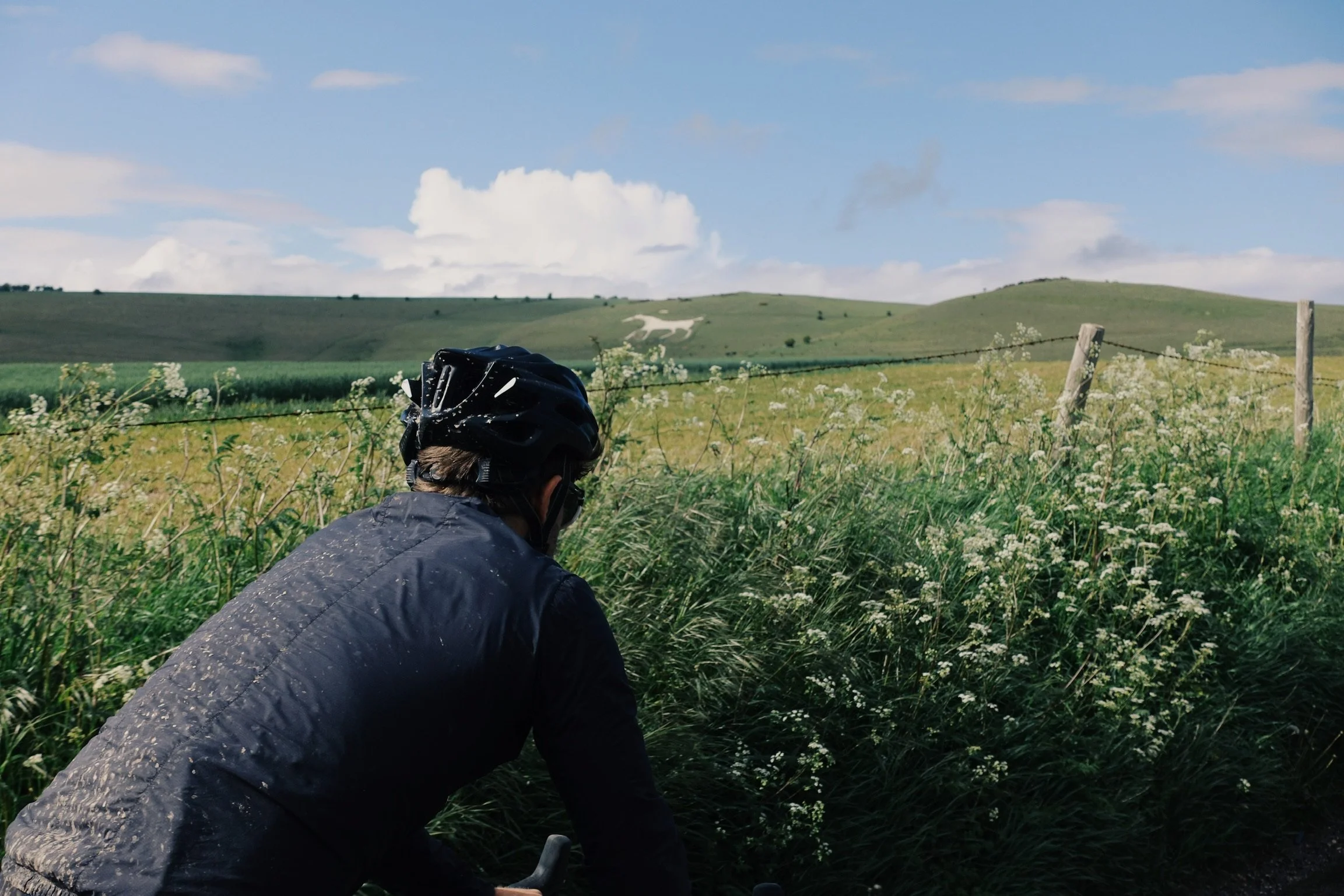 Person wearing a black helmet and dark jacket riding a bicycle through a grassy countryside with green rolling hills and cloudy sky in the background.