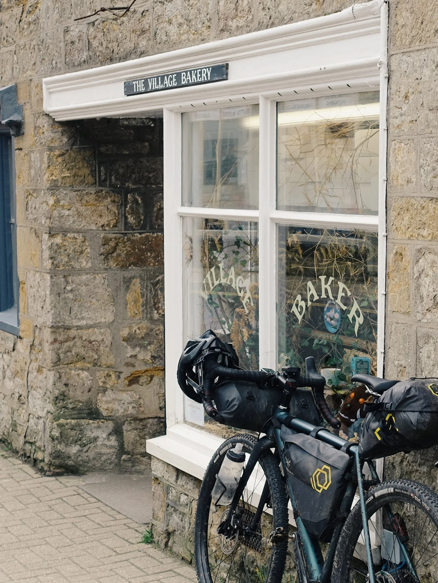 A black bicycle with gear and water bottle parked outside a bakery called 'The Village Bakery' in a stone building. The bakery has a white-framed window with the words 'Village Bakery' painted on the glass.