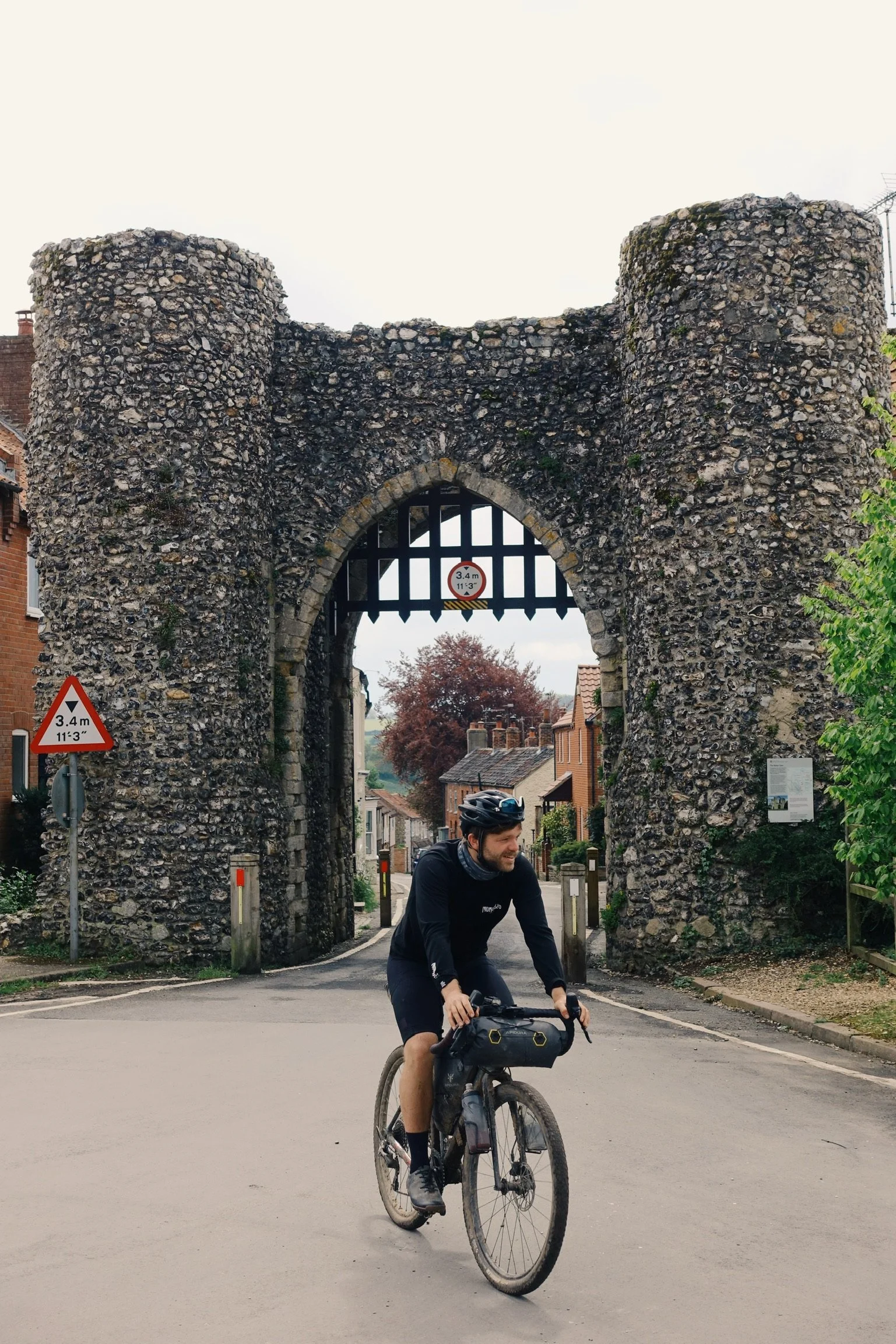 A man riding a bicycle through a historic stone archway in a small town, with trees and houses in the background.