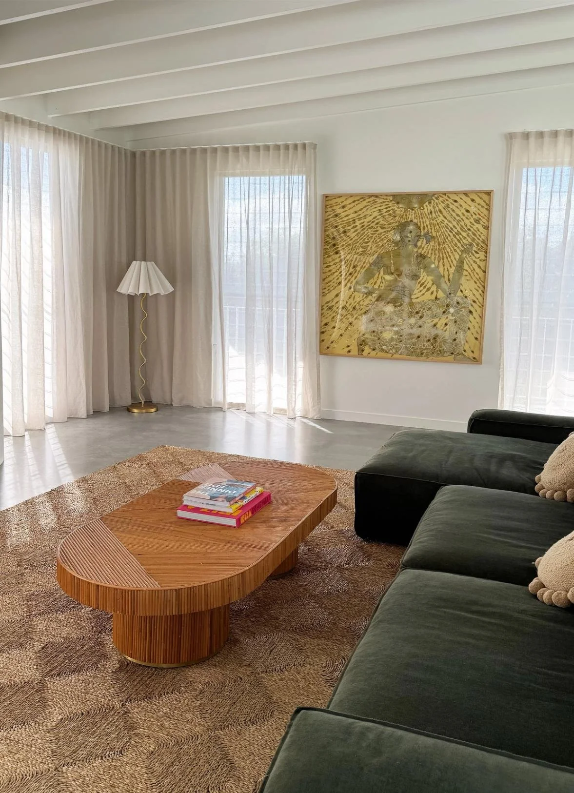 Living room with large windows and sheer curtains, a green sectional sofa with pillows, a wooden coffee table with books, a patterned area rug, a white floor lamp, and a large framed artwork on the wall