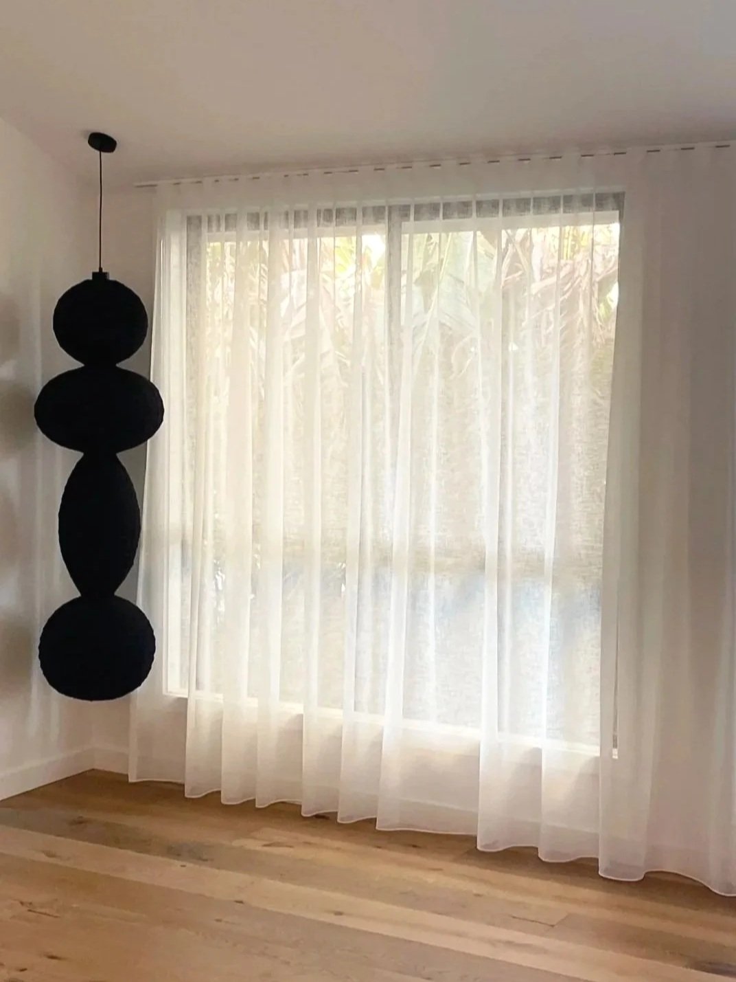 A room with white sheer curtains on a large window and a hanging lamp with three black spherical shades.