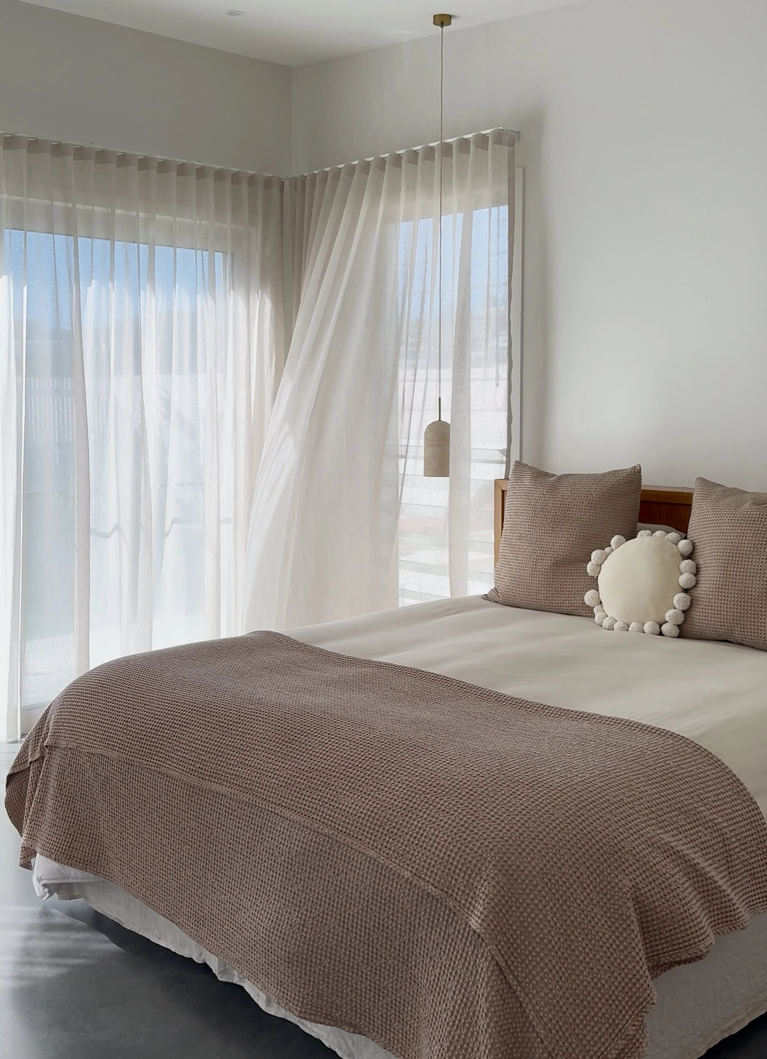 A bedroom featuring a bed with a brown textured bedspread, beige pillows including one with pom-pom trim, and sheer curtains on windows with a view of the outdoors. There is a hanging light fixture near the window.