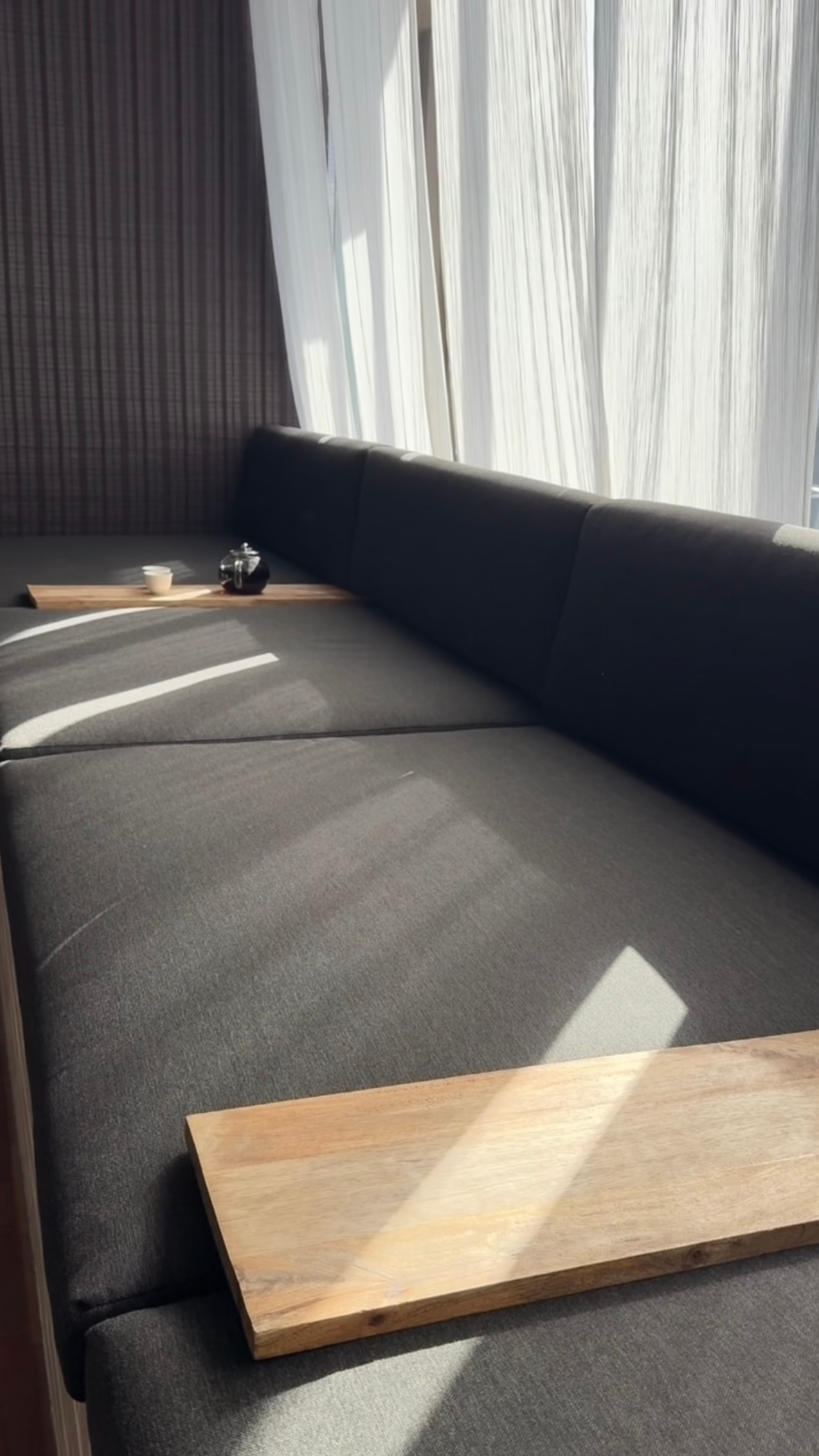A dark gray sofa with a wooden coffee table placed on it. The coffee table is made of light-colored wood and has a simple rectangular shape. In the background, there are large white curtains, with sunlight casting shadows on the sofa.