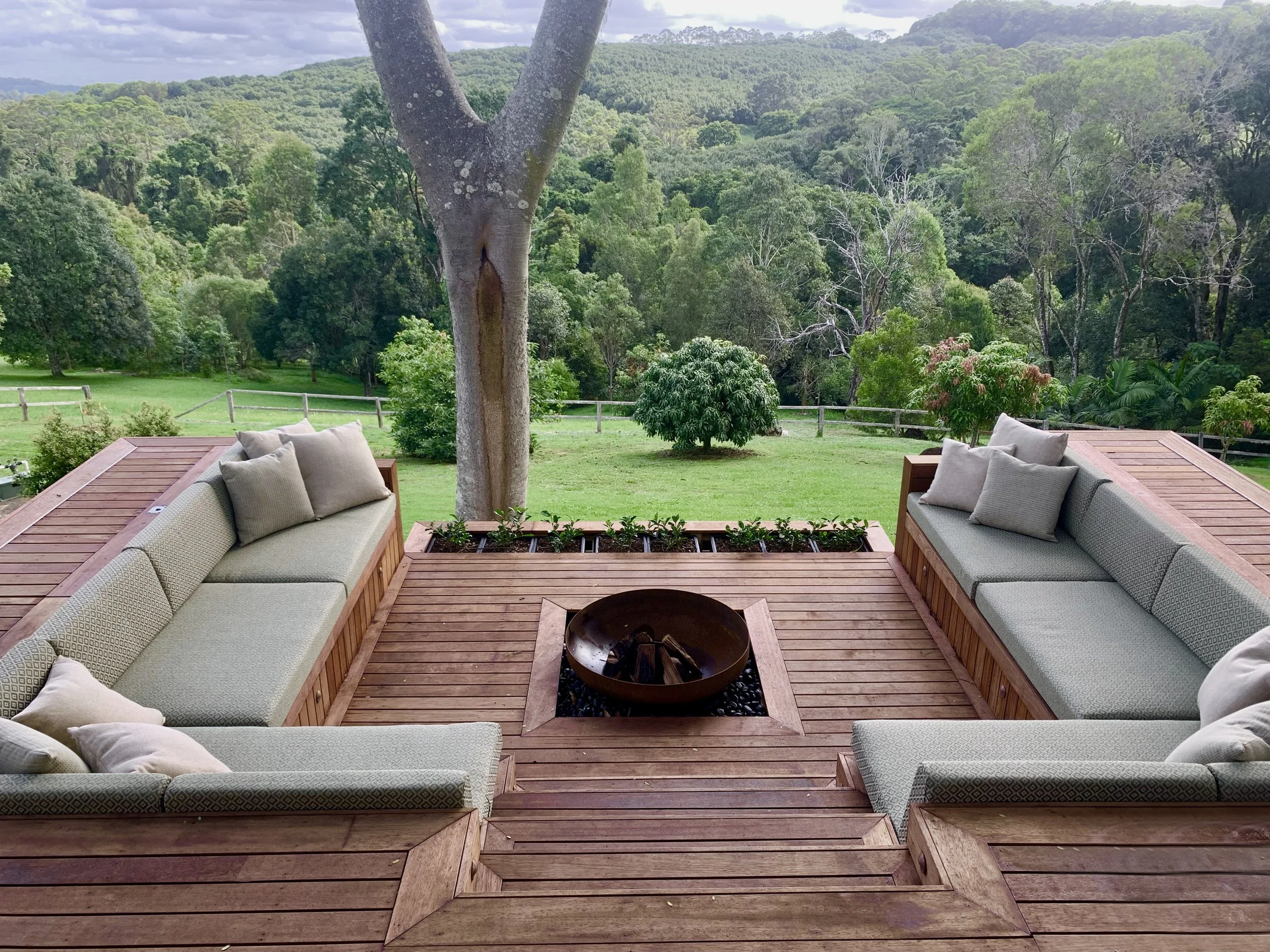 Outdoor deck with built-in seating surrounding a fire pit, overlooking a green backyard with trees and rolling hills in the background.