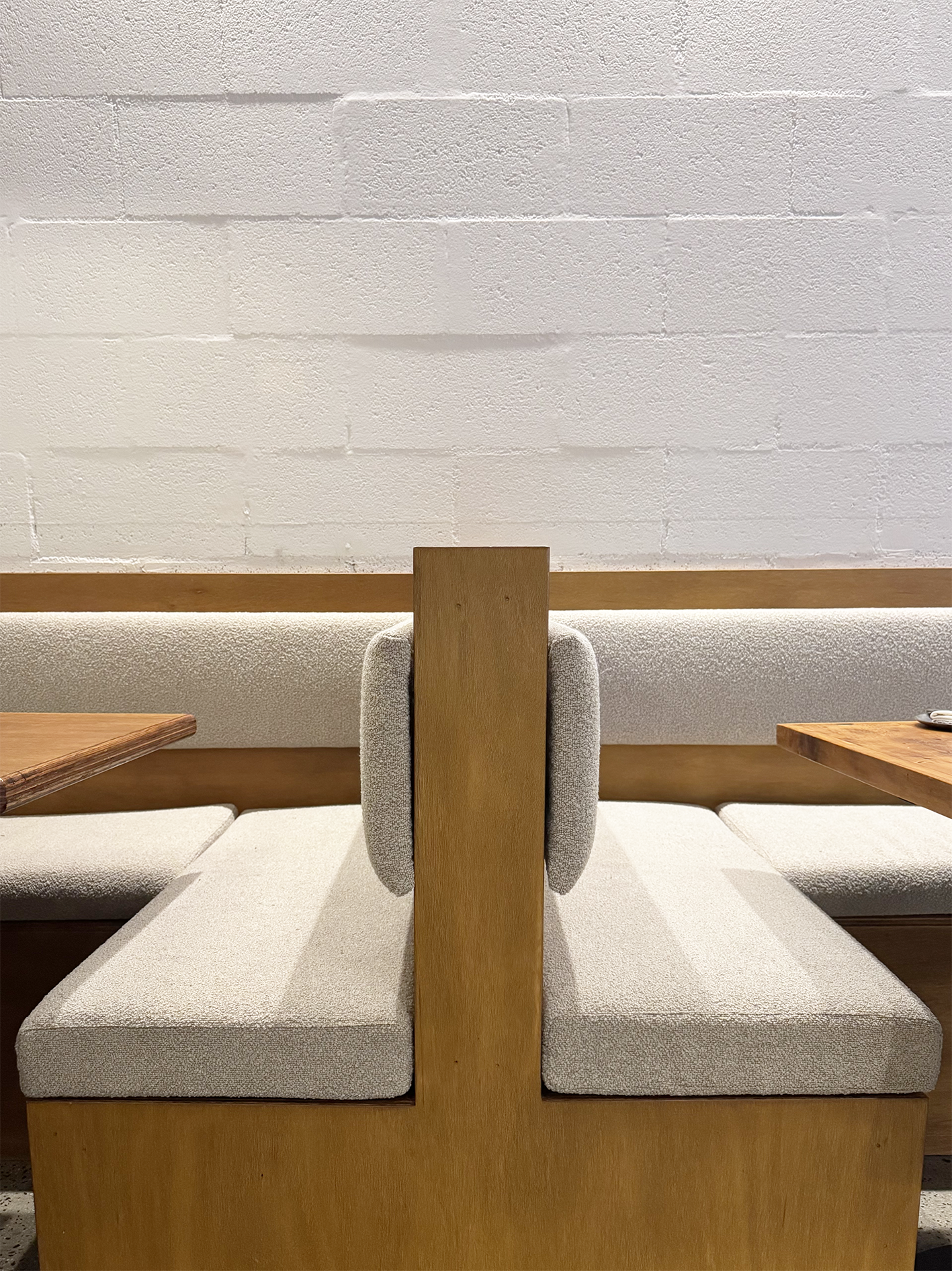 A beige cushioned seating area with a wooden divider and a chair with a light gray backrest in front of a plain white textured wall.