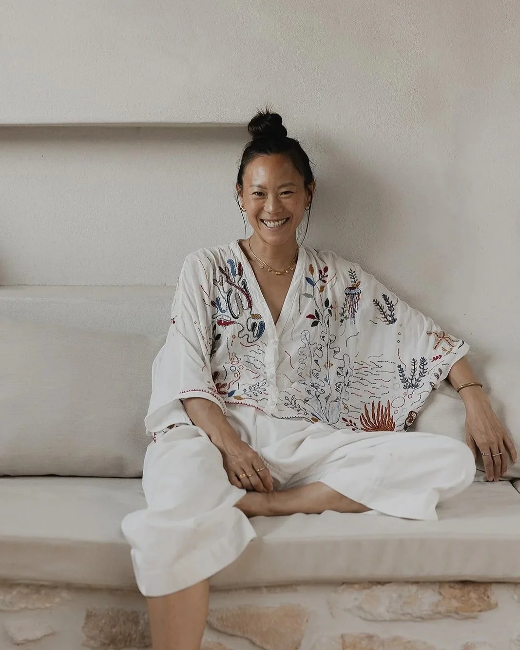 A smiling woman with dark hair in a bun, wearing a white embroidered blouse and white pants, sitting cross-legged on a beige couch against a plain white wall.