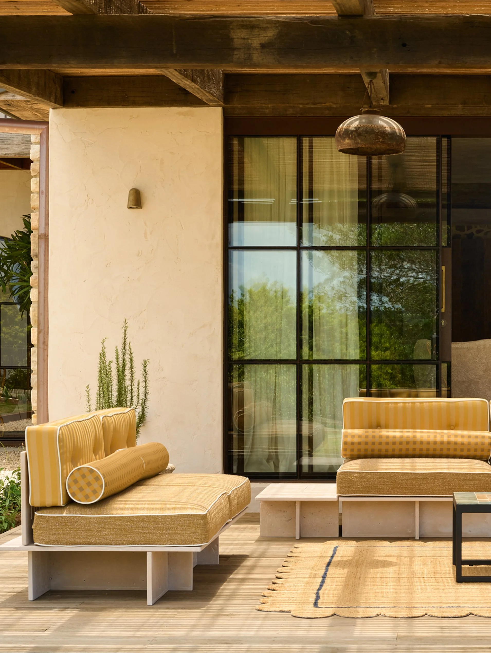 Outdoor patio area with yellow cushioned seating, a rug, a white table, a plant, and large glass windows reflecting trees.