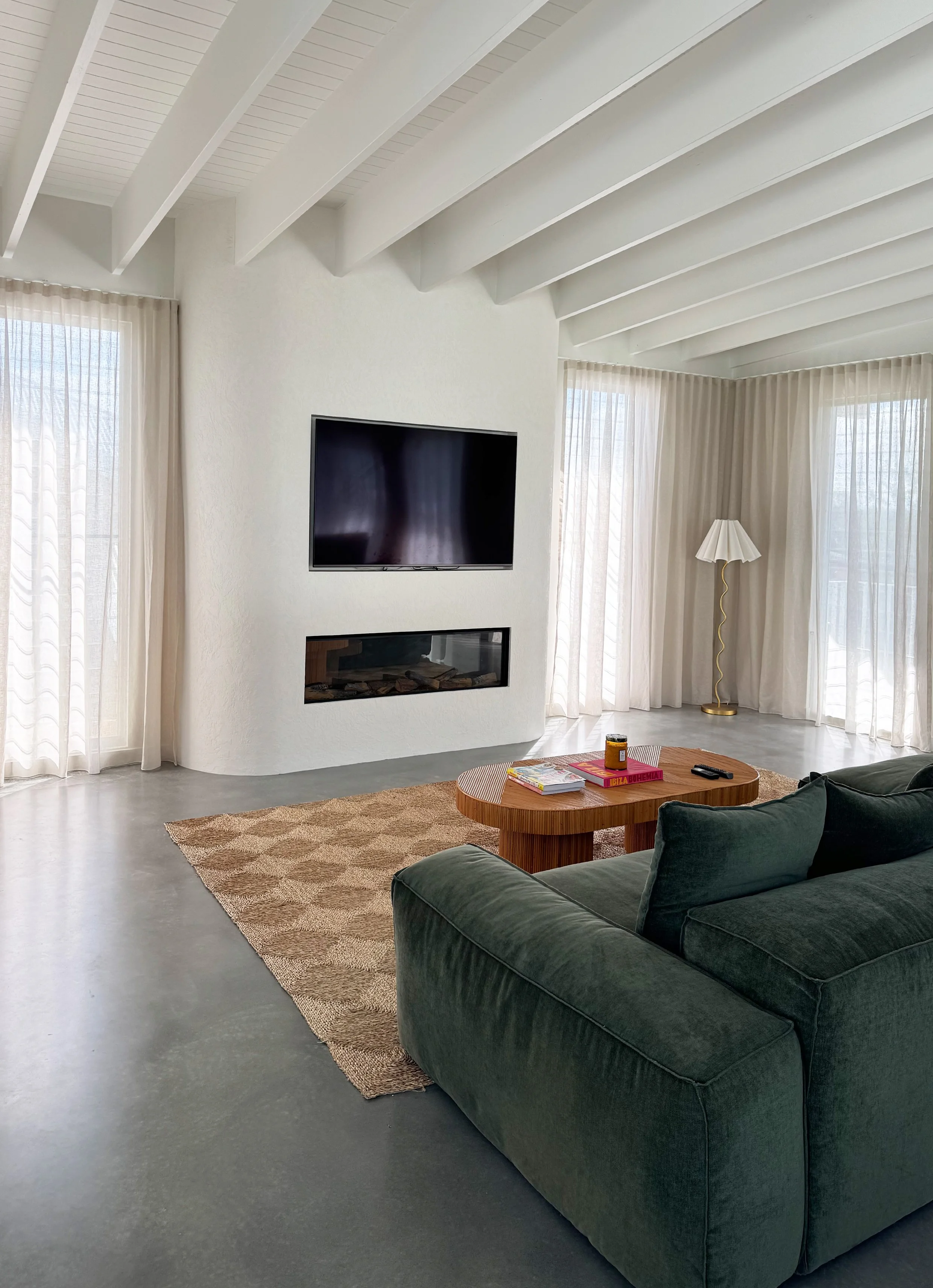 Living room with green velvet sofa, wooden coffee table with magazines and candles, beige rug, TV mounted on white wall, fireplace below, large windows with sheer curtains, and a tall floor lamp with a white shade and gold stand.