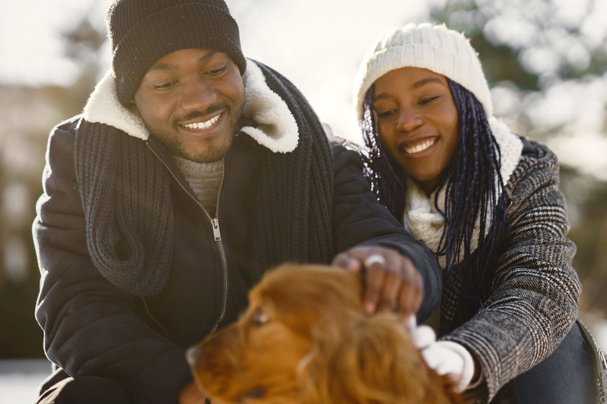 Two adults in winter clothing pet a golden-brown dog outdoors.