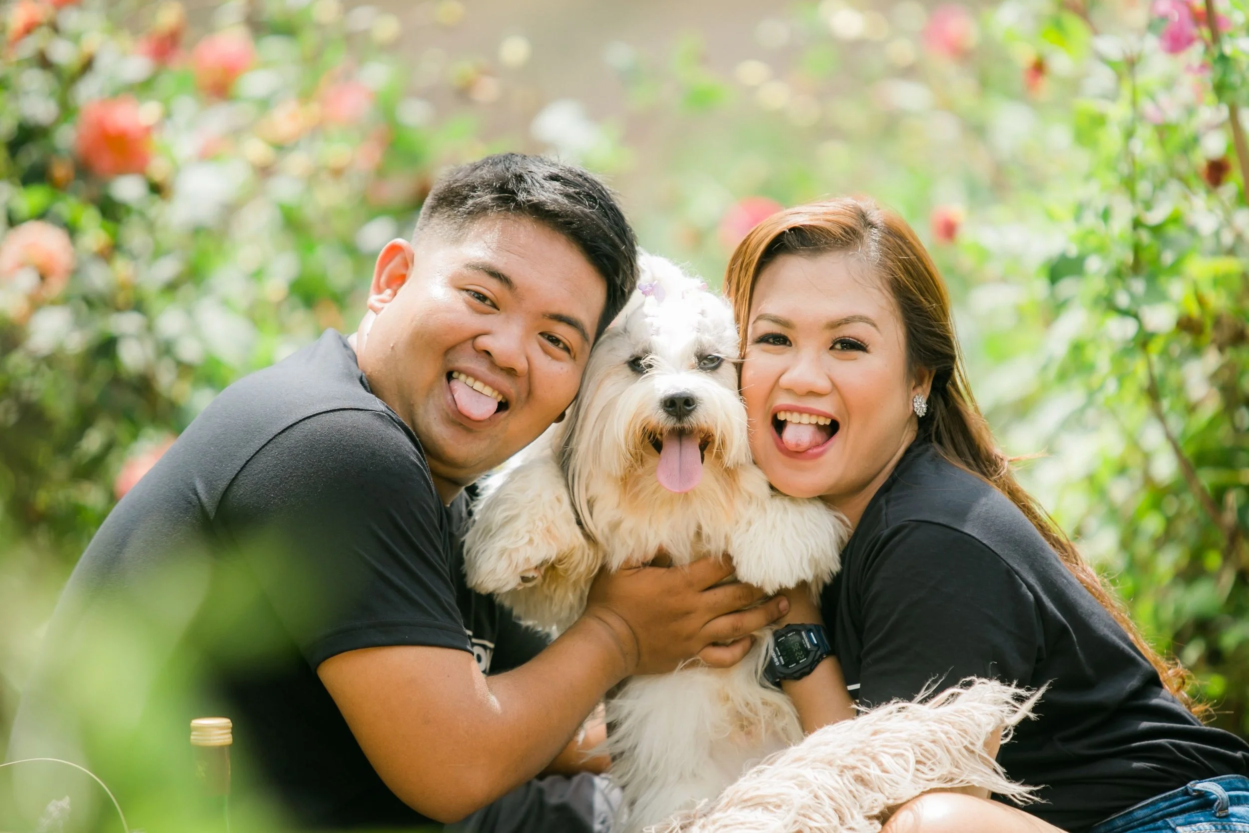 Two smiling people hold a fluffy white dog in a garden.