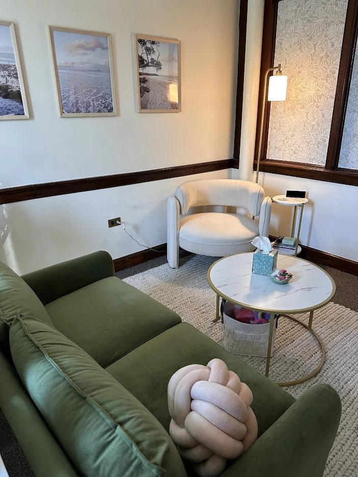 A cozy living room with a green sofa, a white armchair, and a round coffee table with tissues and a small bowl. A tall standing lamp is beside the armchair. Three framed landscape pictures hang on the wall, and a window with frosted patterned glass is nearby. A small side table with books and a phone is next to the armchair.