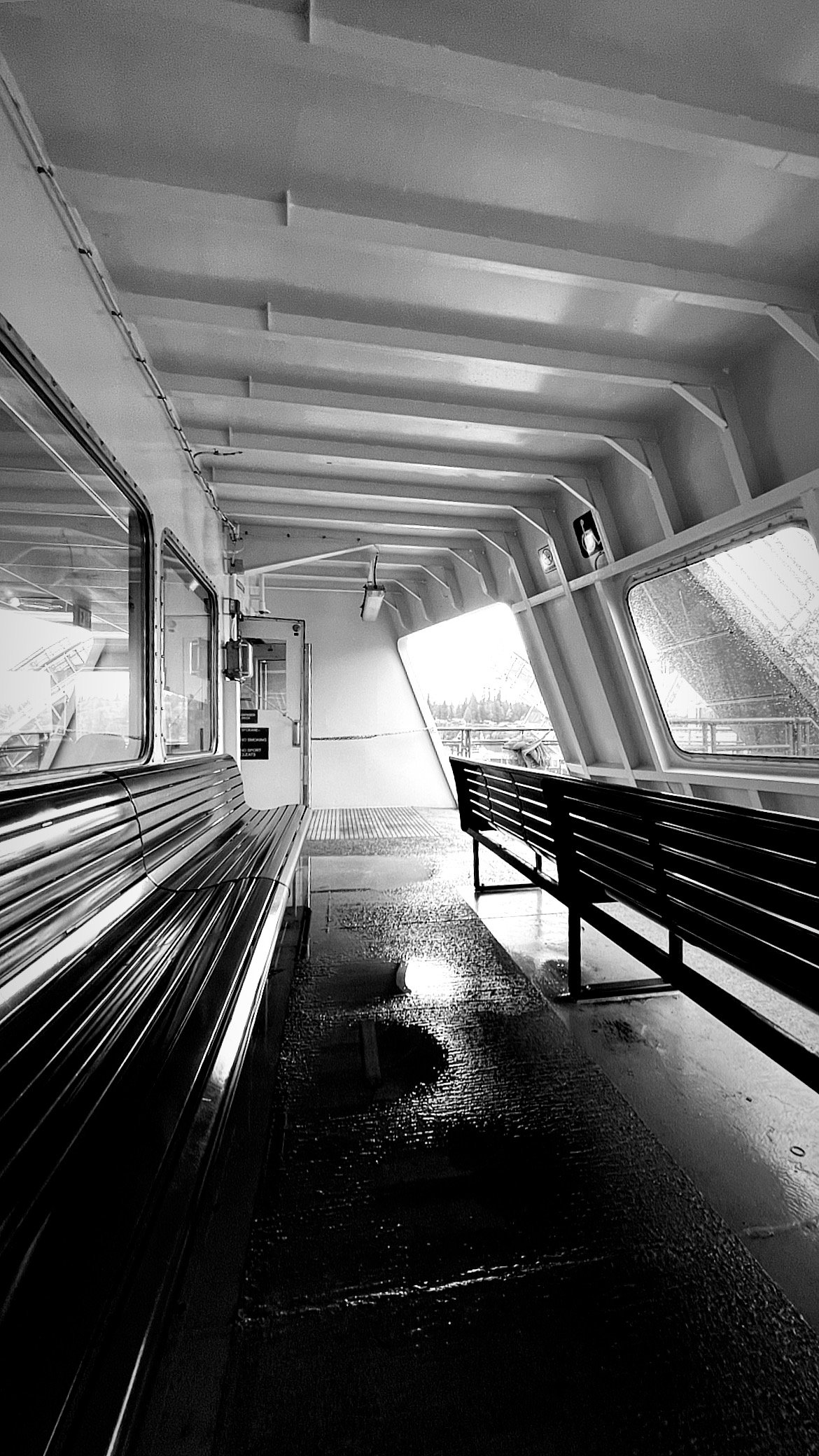 Black and white photo inside the passenger area of a ferry with empty benches and a wet deck, showing windows overlooking a rainy day at sea, capturing the quiet stillness of travel between two shores.