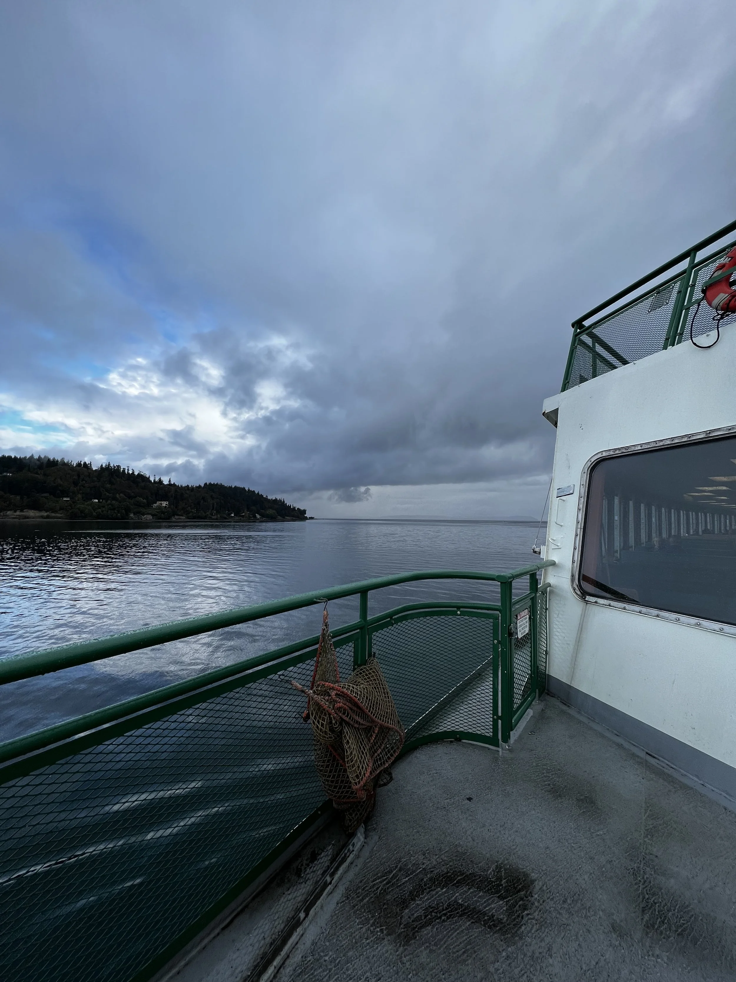 View from the ferry deck showing a lifebuoy attached to the green railing, wet floor beneath, and a cloudy sky above meeting the calm sea on the horizon, evoking a feeling of tranquil passage and natural harmony