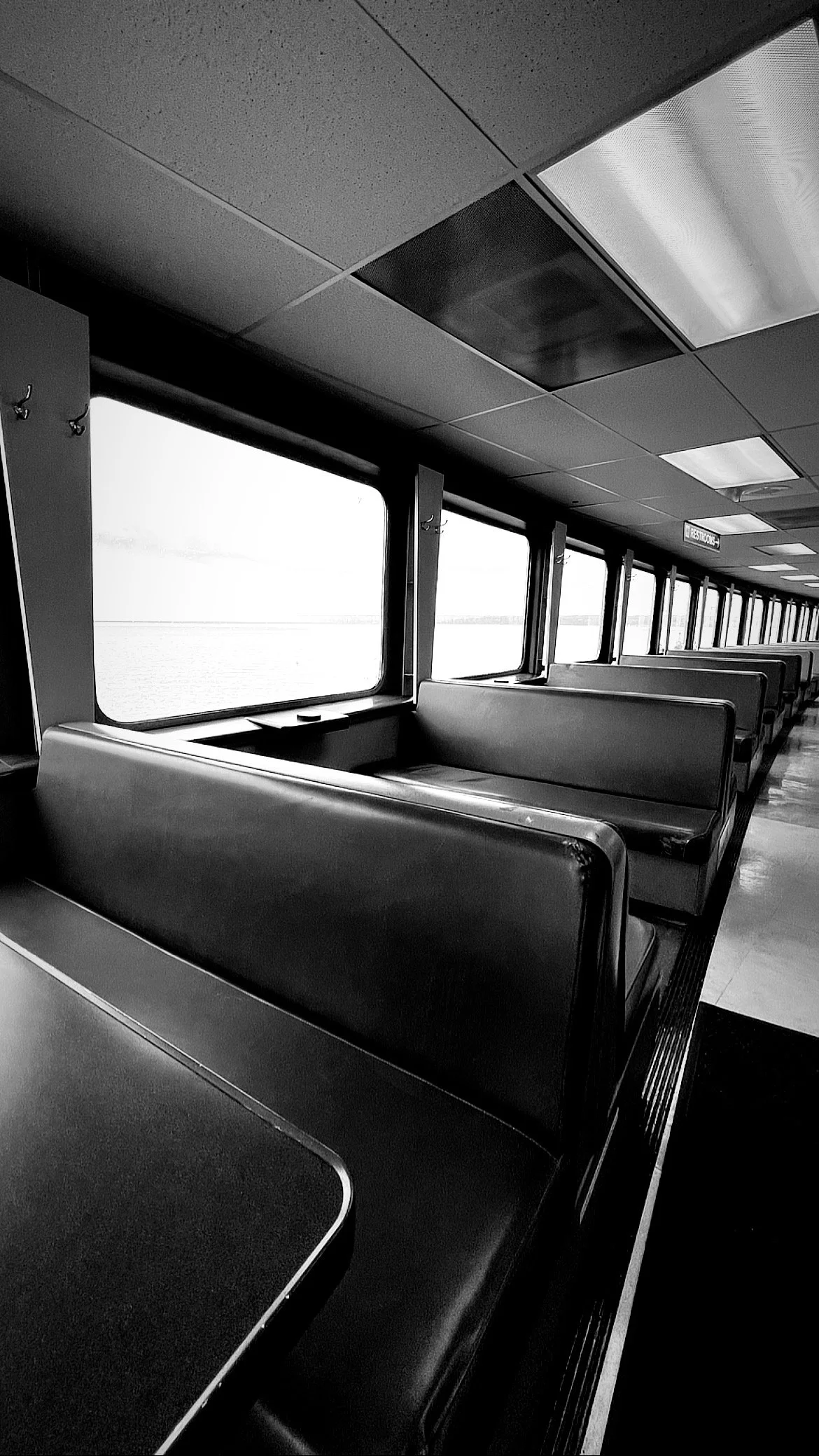 Empty passenger seating inside a ferry with large windows showcasing a dramatic view of the overcast sky and distant shore, all in monochrome.