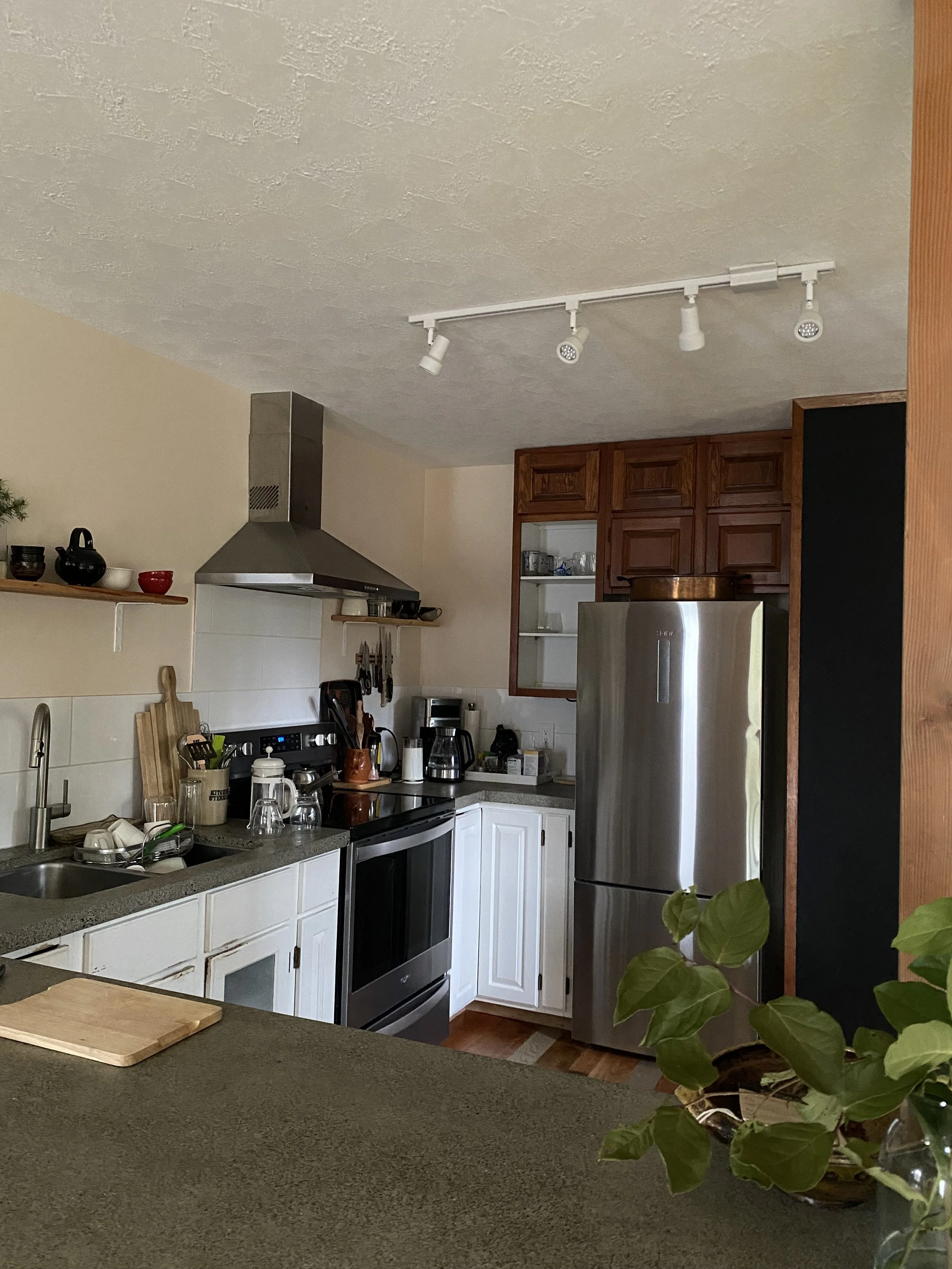 A kitchen with a silver refrigerator, white cabinetry, a stove, and a gray countertop. There are pots, jars, and kitchen utensils on the counter and a wooden cutting board. A plant with green leaves is in the foreground.