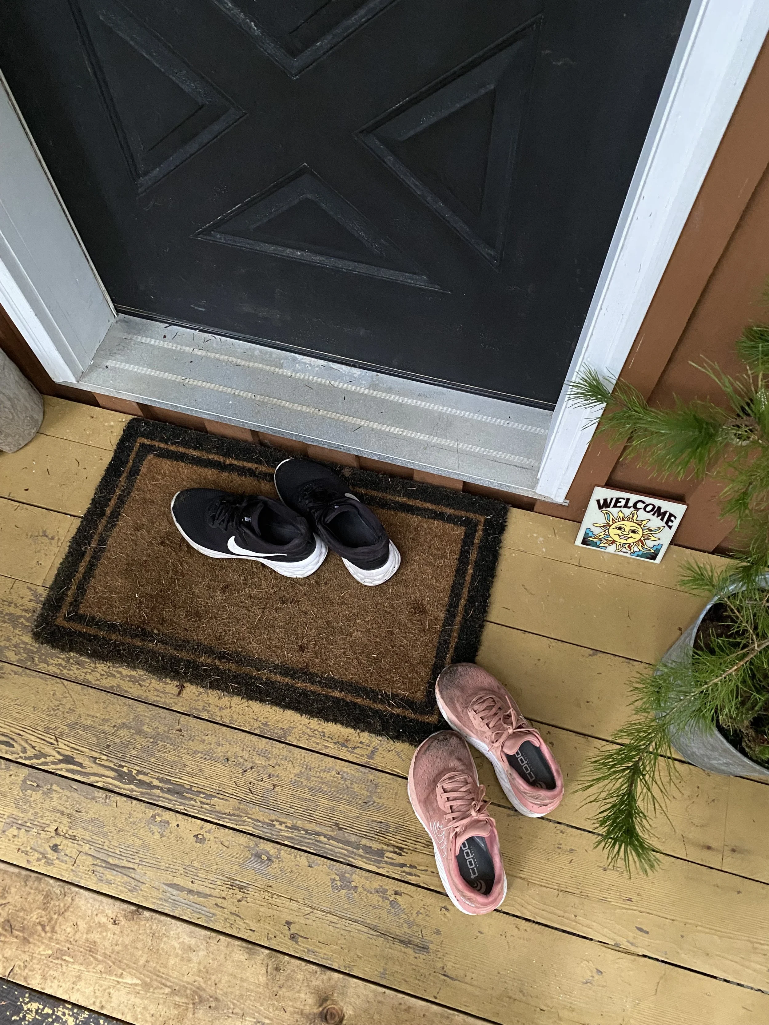 View of a front door area with two pairs of shoes, one black and white athletic shoes and one pink athletic shoes, placed on a brown doormat in front of a black door. To the right of the door, a small welcome sign and a potted plant are visible on a 
